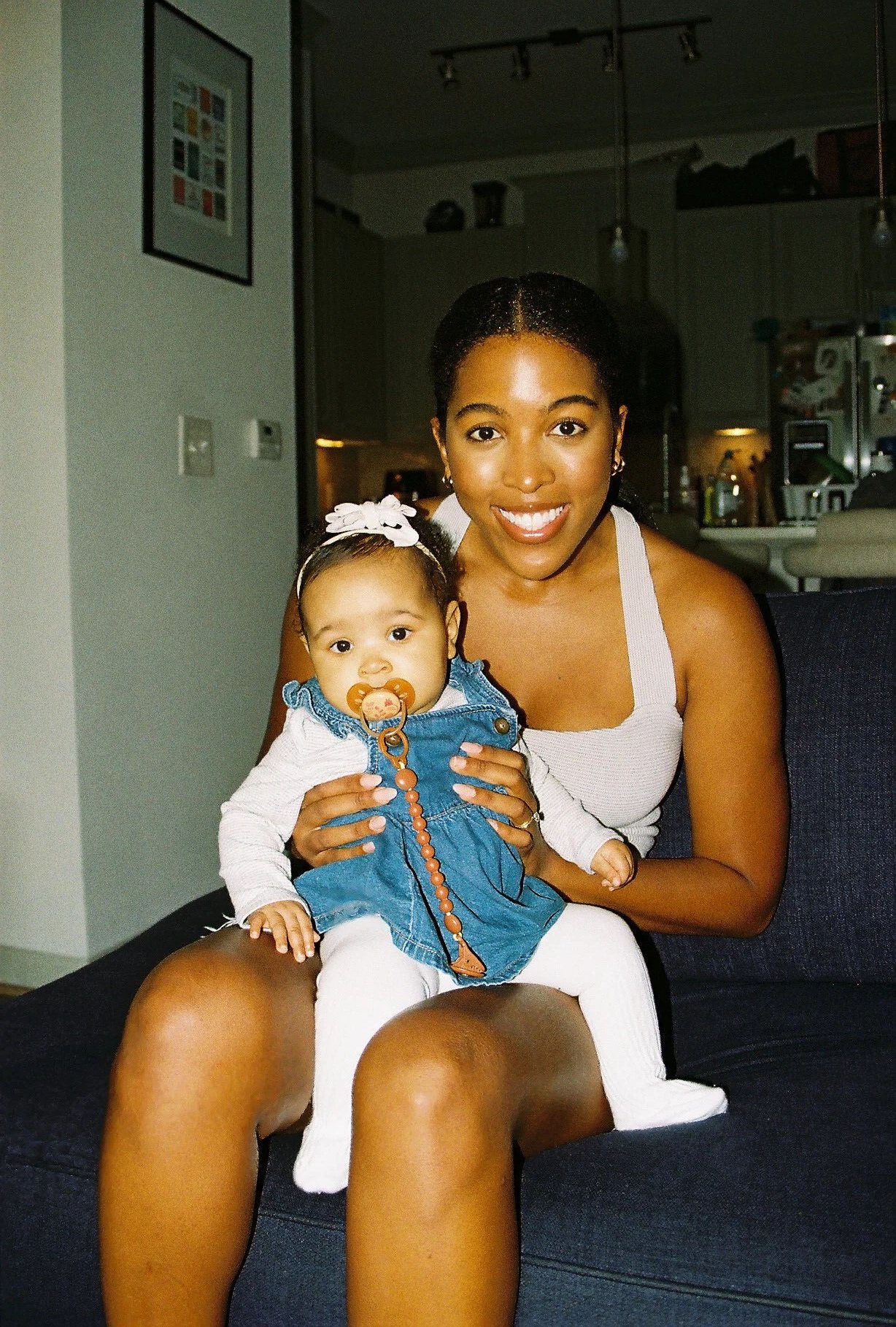 A smiling woman holding a baby girl with a pacifier, sitting on a dark-colored sofa in a room with a kitchen background.