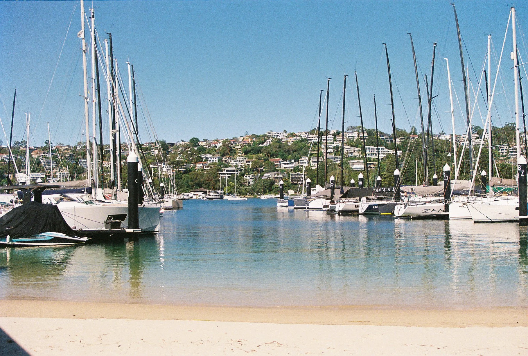 View of a marina with sailboats docked, hills with houses in the background, sandy beach in the foreground, and a clear blue sky.