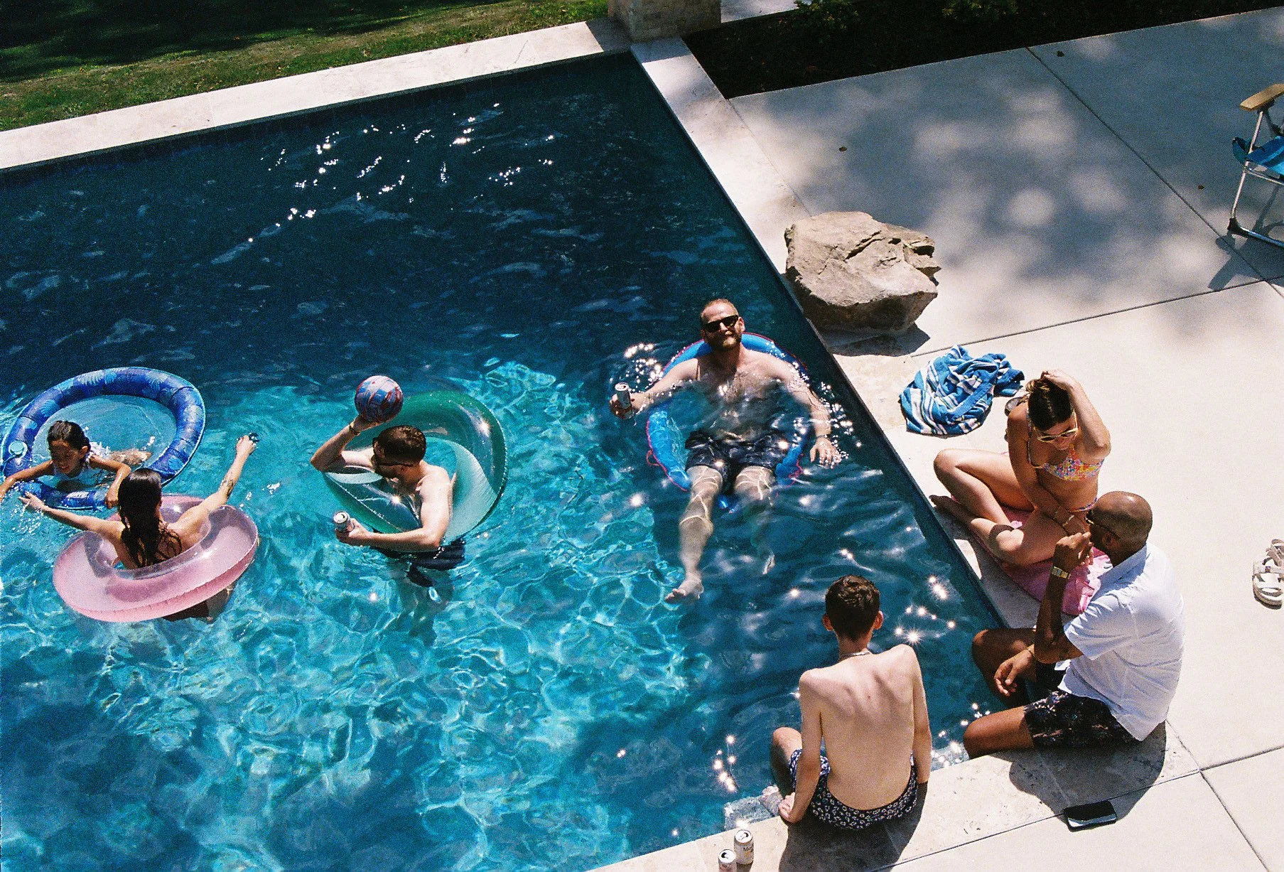 People enjoying a sunny day at a backyard pool, with children and adults swimming, playing, and relaxing on the poolside.