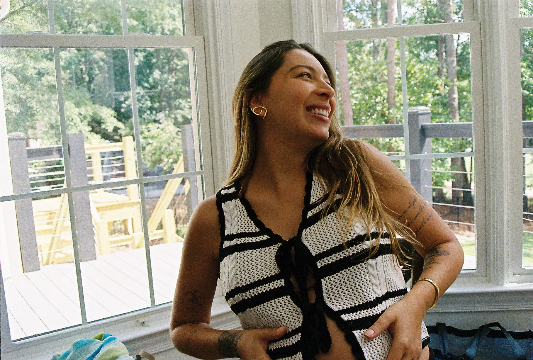 Young woman with long brown hair smiling, wearing a black and white crochet top, standing indoors near large windows with a wooden deck and green trees outside.