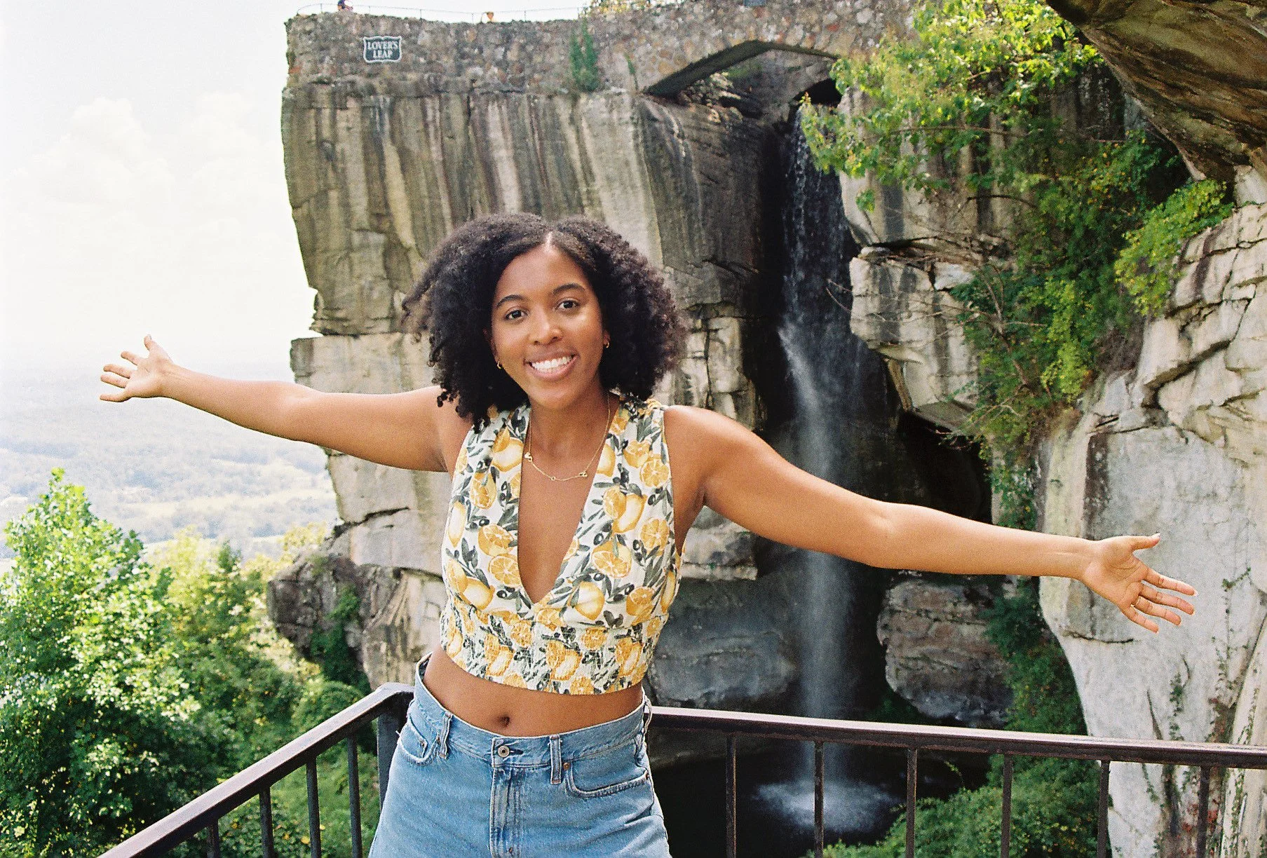 A smiling woman with curly hair in a sleeveless lemon-patterned top and jeans, standing with arms outstretched in front of a waterfall and rocky cliff, with forest in the background.