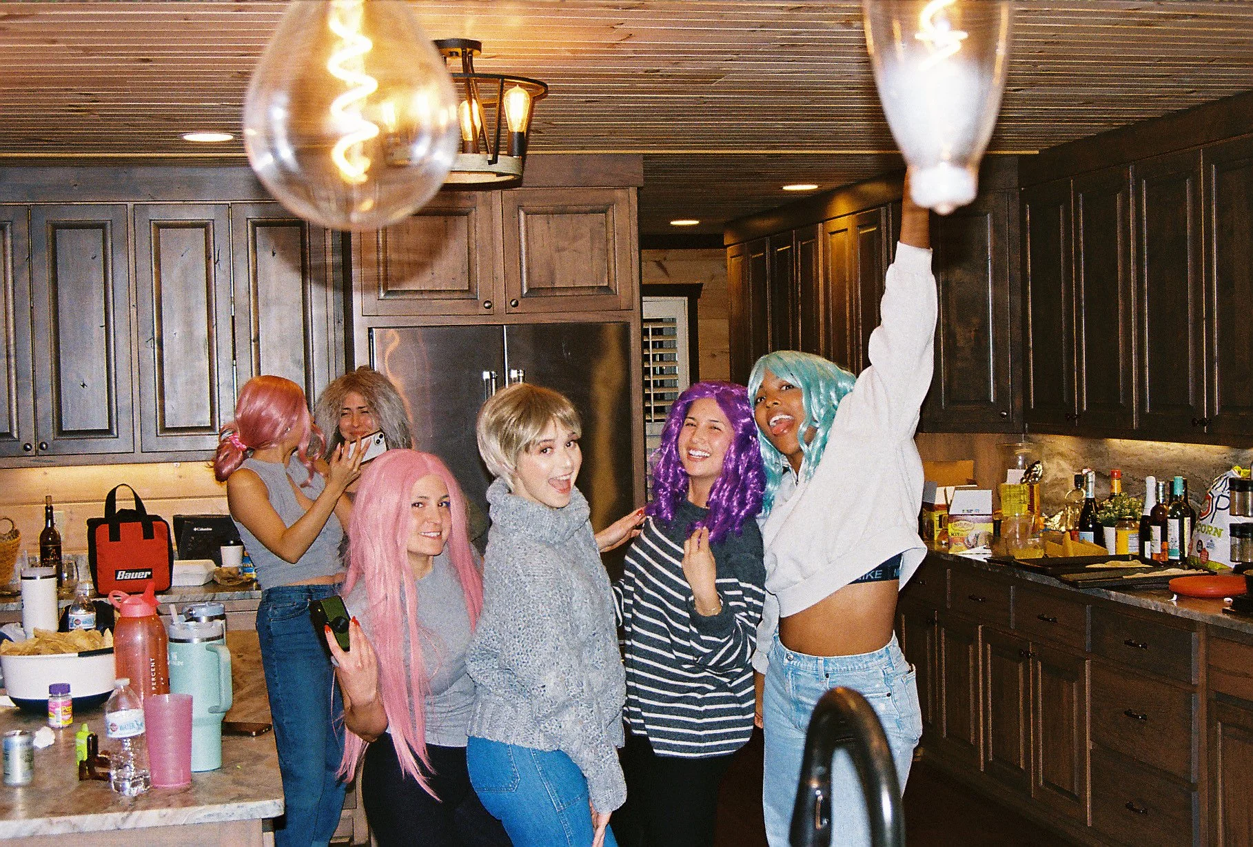 Group of five young women with colorful hair celebrating in a kitchen. One woman is hanging a light bulb, and others are smiling or taking photos.