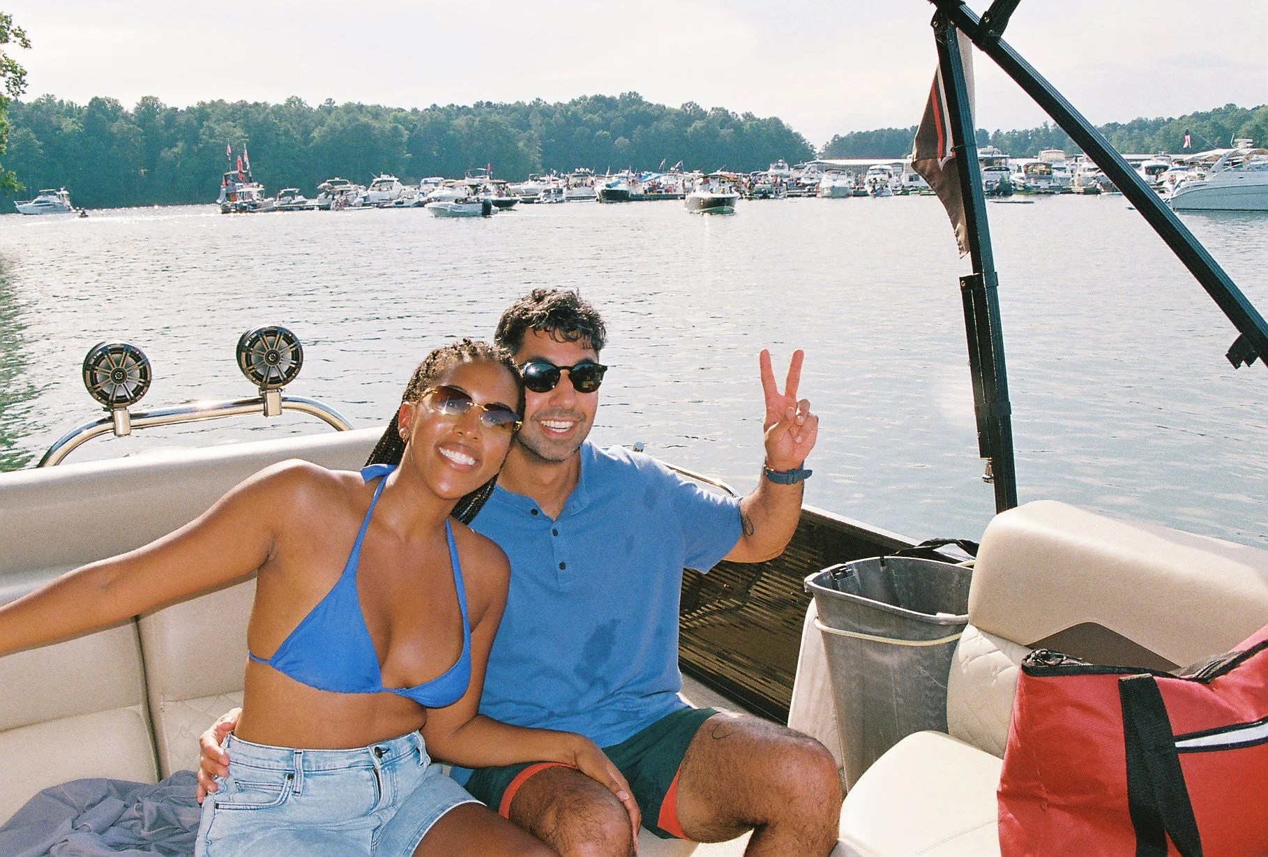 A smiling woman and man enjoying a boat ride on a lake, with multiple boats and a tree-lined shoreline in the background. The woman is wearing sunglasses and a blue bikini top, while the man is wearing sunglasses and a blue shirt, making a peace sign