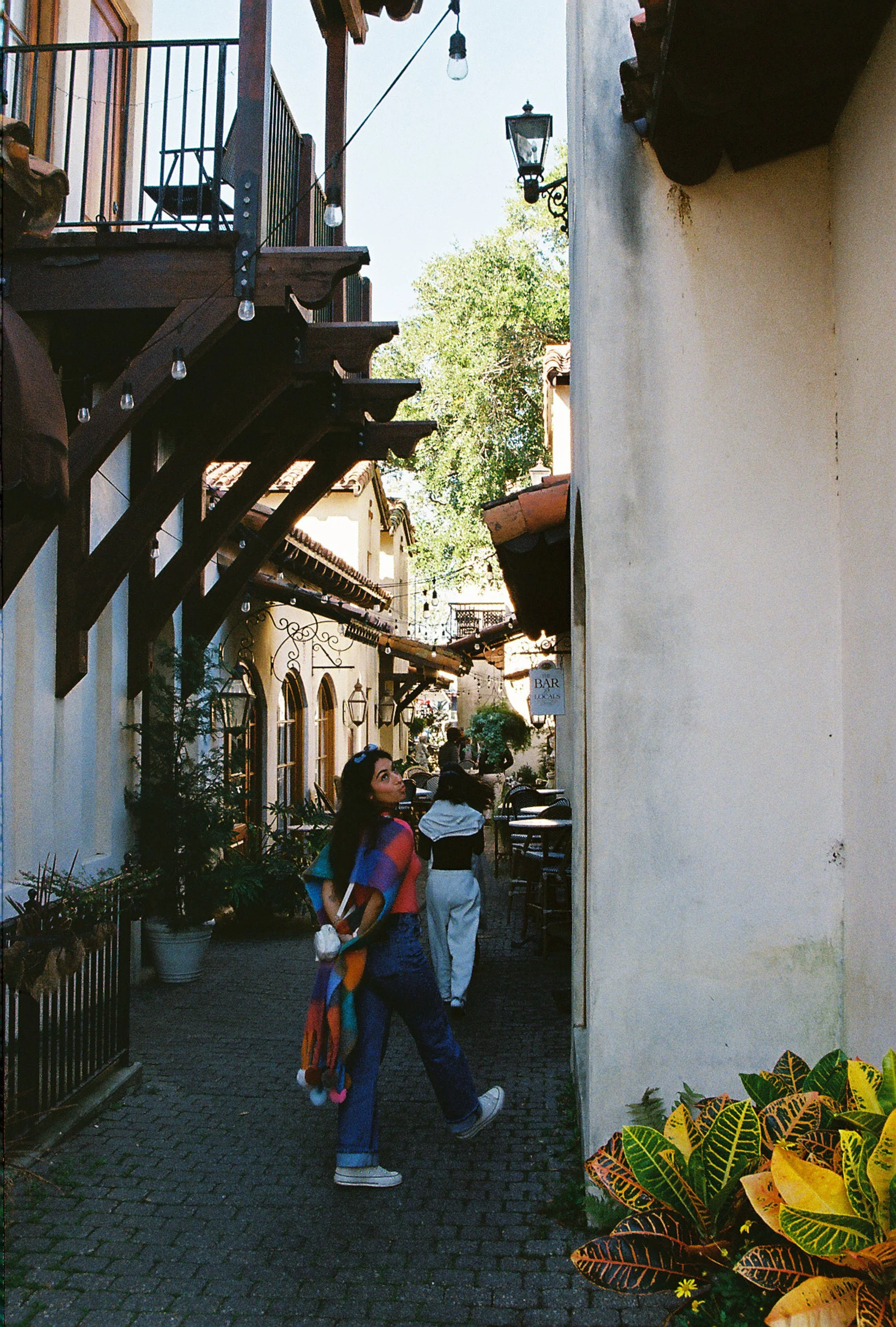 A woman in colorful attire walking along a narrow outdoor street with shops and potted plants, under a clear sky.