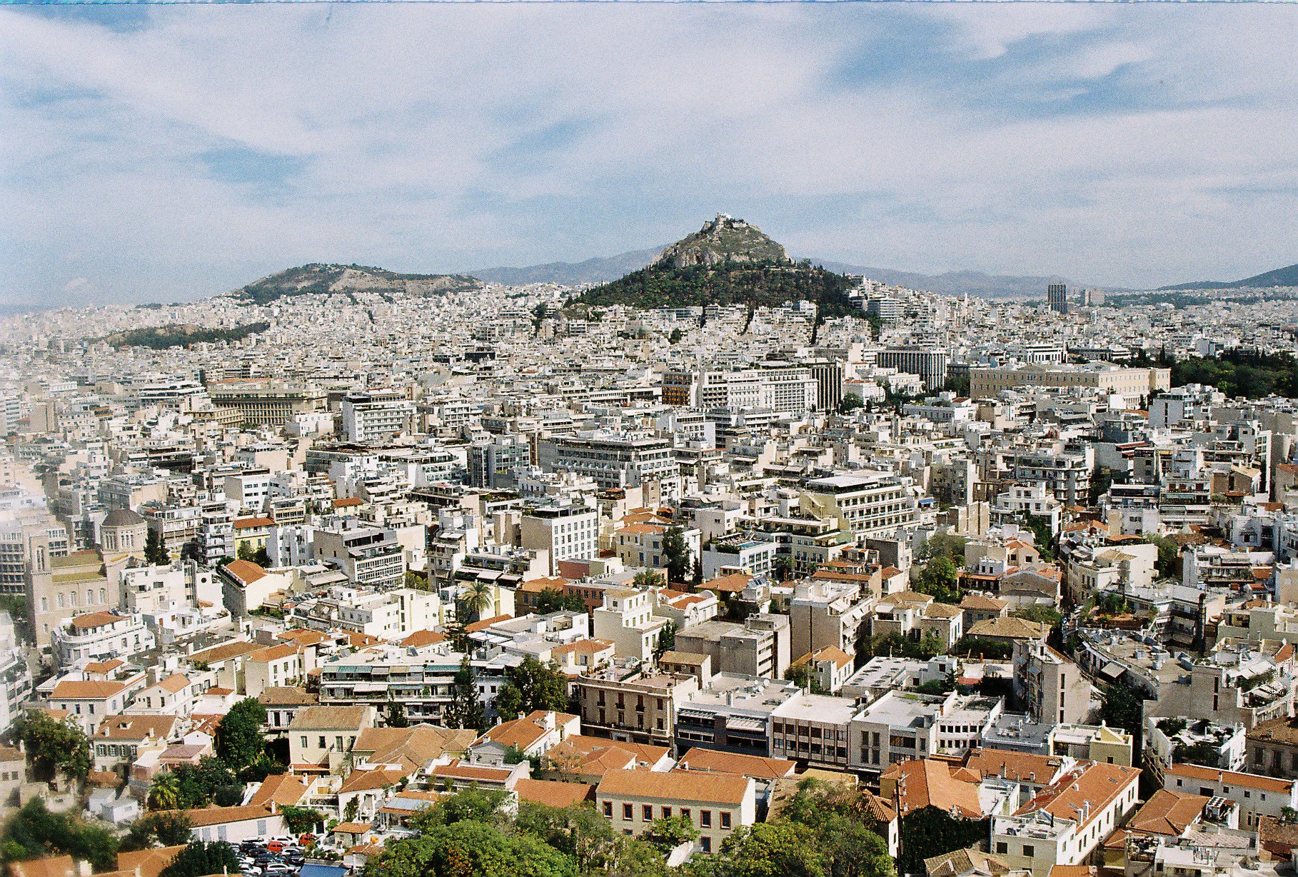 A wide cityscape of Athens, Greece with Mount Lycabettus in the background and densely packed white and beige buildings in the foreground.