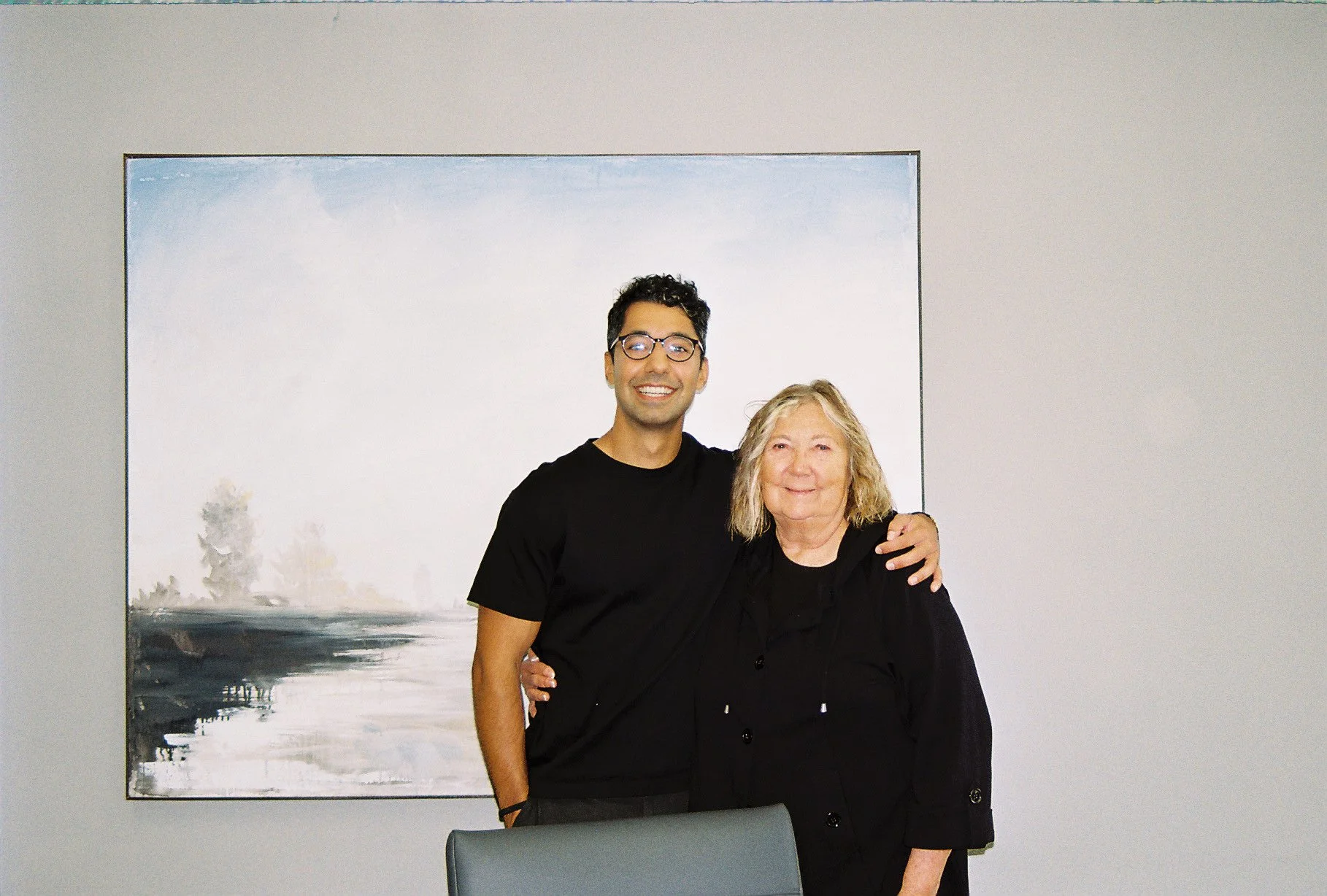 Two people, a man and a woman, standing close together and smiling, with the man having his arm around the woman's shoulder, in front of a painting on a plain wall.