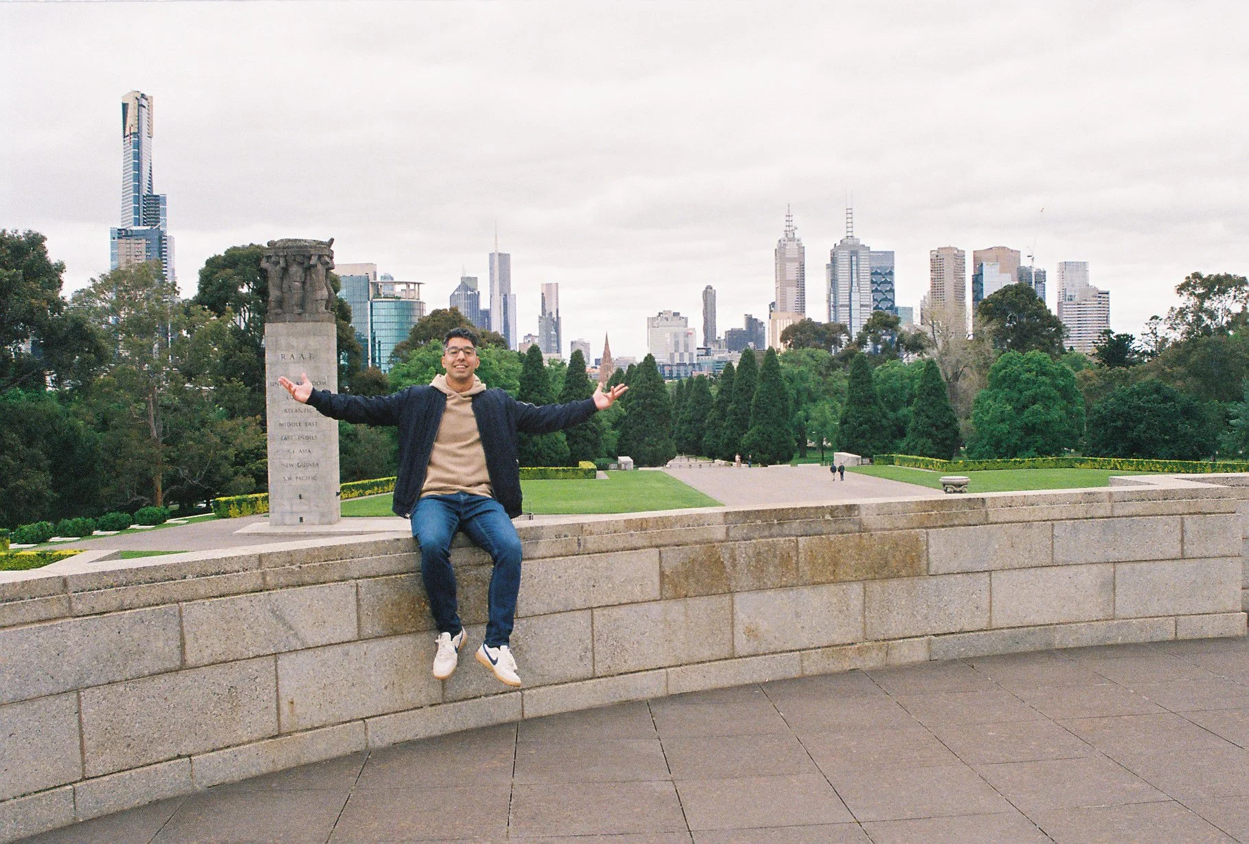 A young man sits on a stone ledge with arms outstretched, smiling, in a city park with a view of tall skyscrapers in the background.