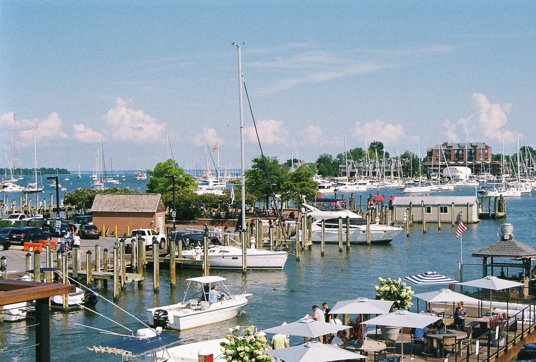 A marina filled with sailboats and motorboats docked at wooden piers, with people dining under umbrellas on an outdoor deck, and a parking lot nearby with cars parked, under a partly cloudy sky.