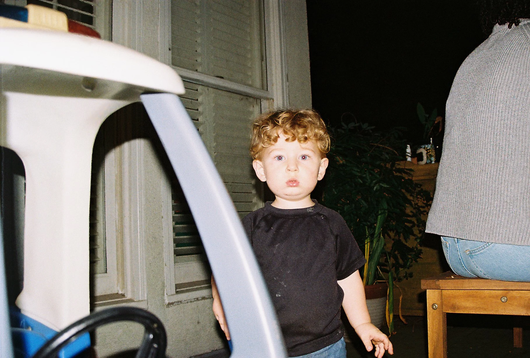 A young boy with curly red hair and blue eyes standing outside at night near a window and a person sitting on a wooden bench. The boy is wearing a black t-shirt and appears to be surprised or curious.