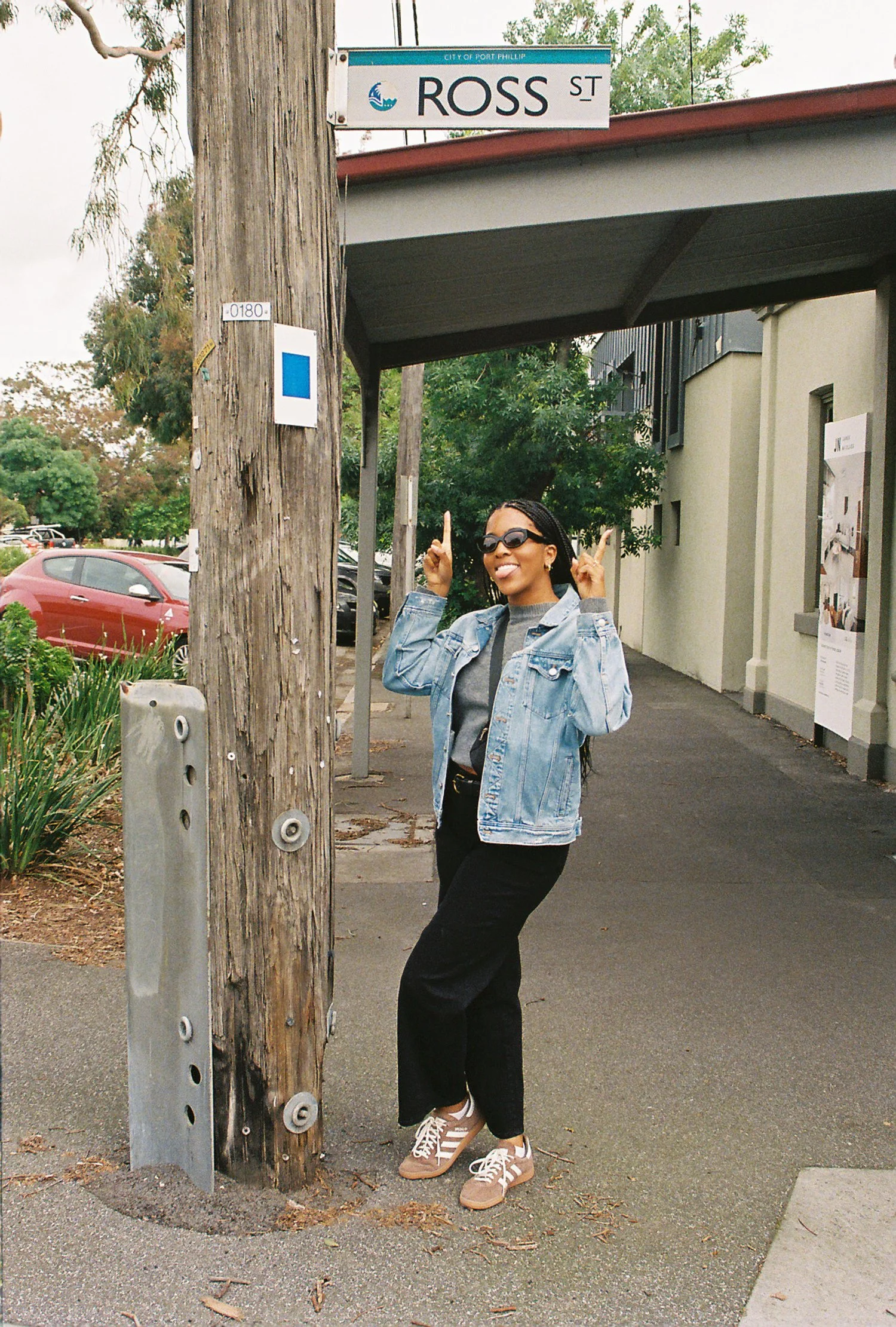A woman with short black hair and sunglasses standing on a sidewalk next to a wooden utility pole, pointing upwards with both hands, smiling, wearing a denim jacket, gray shirt, black pants, and beige sneakers. In the background, there are trees, par