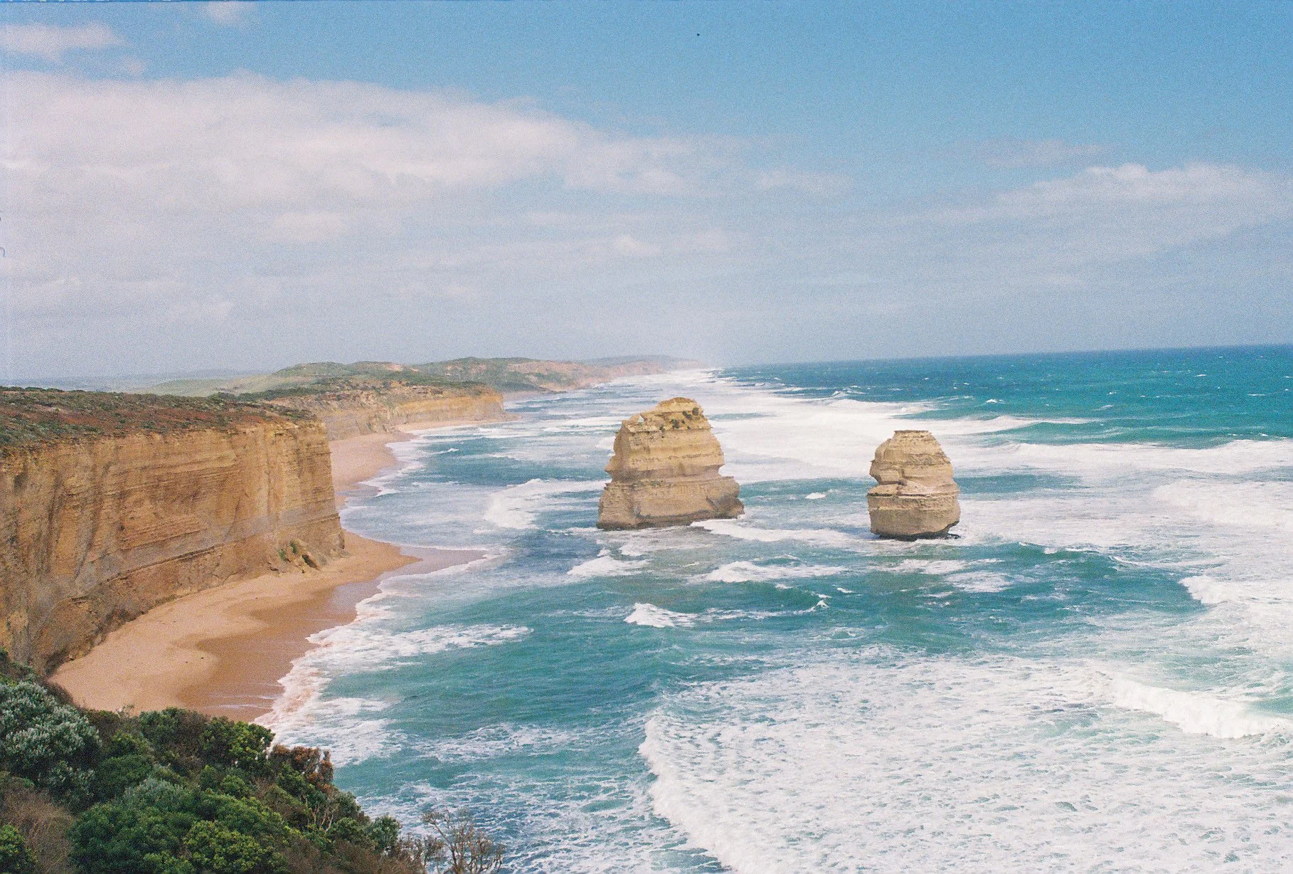 Scenic view of a rugged coastline with tall cliffs and two large sea stacks rising from the ocean near a sandy beach. The ocean waves are crashing against the shore and the sky is partly cloudy.