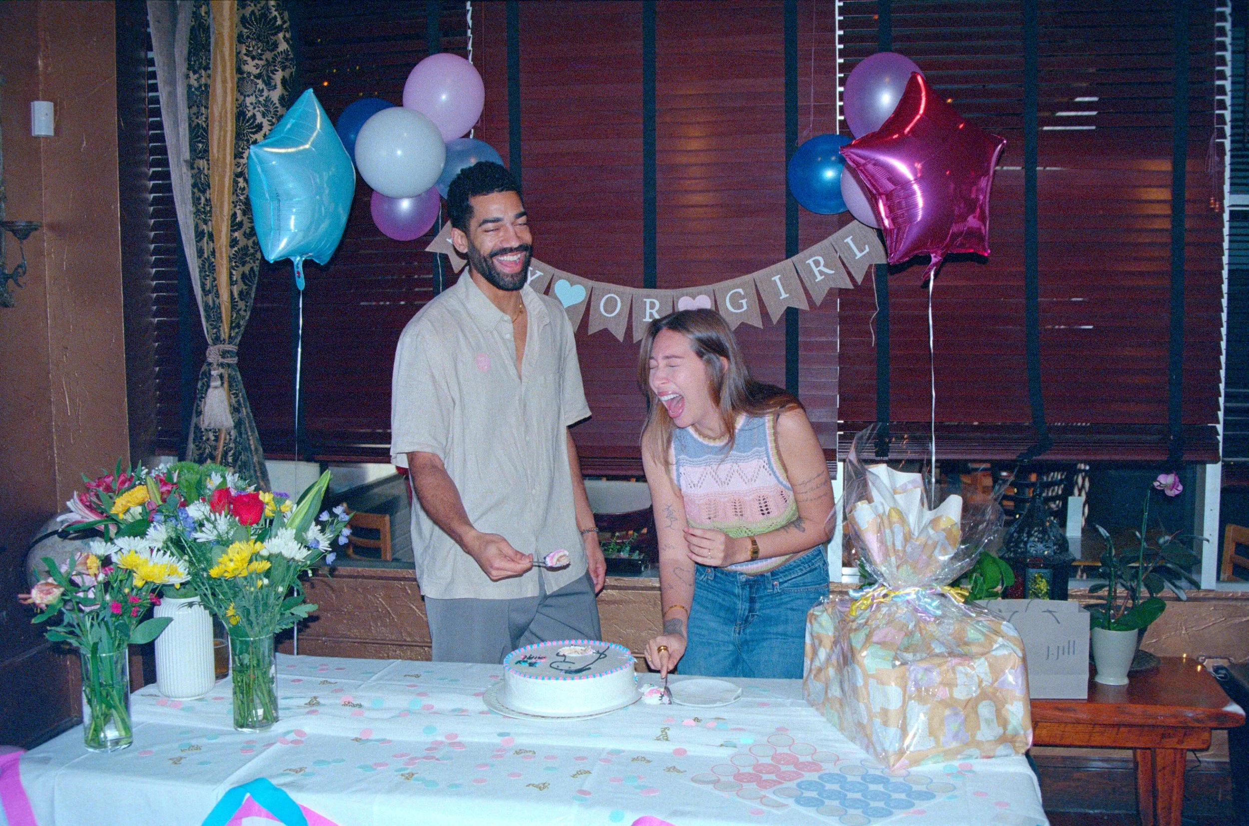 A man and a woman celebrating a birthday with a cake, balloons, flowers, and gifts. The woman is cutting the cake while both are smiling and laughing. A banner in the background reads 'Your Girl'.