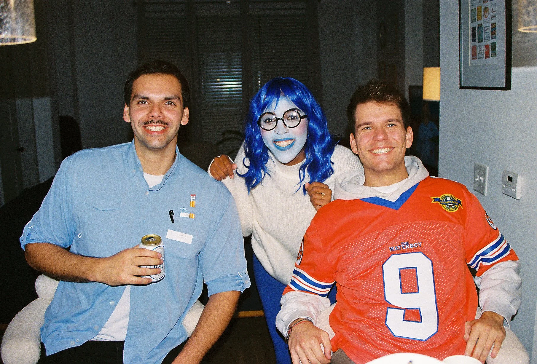Three people smiling indoors, one person with bright blue hair and face paint, wearing large round glasses, and two young men, one in a light blue shirt holding a soda can, and the other in an orange hockey jersey with the number nine.