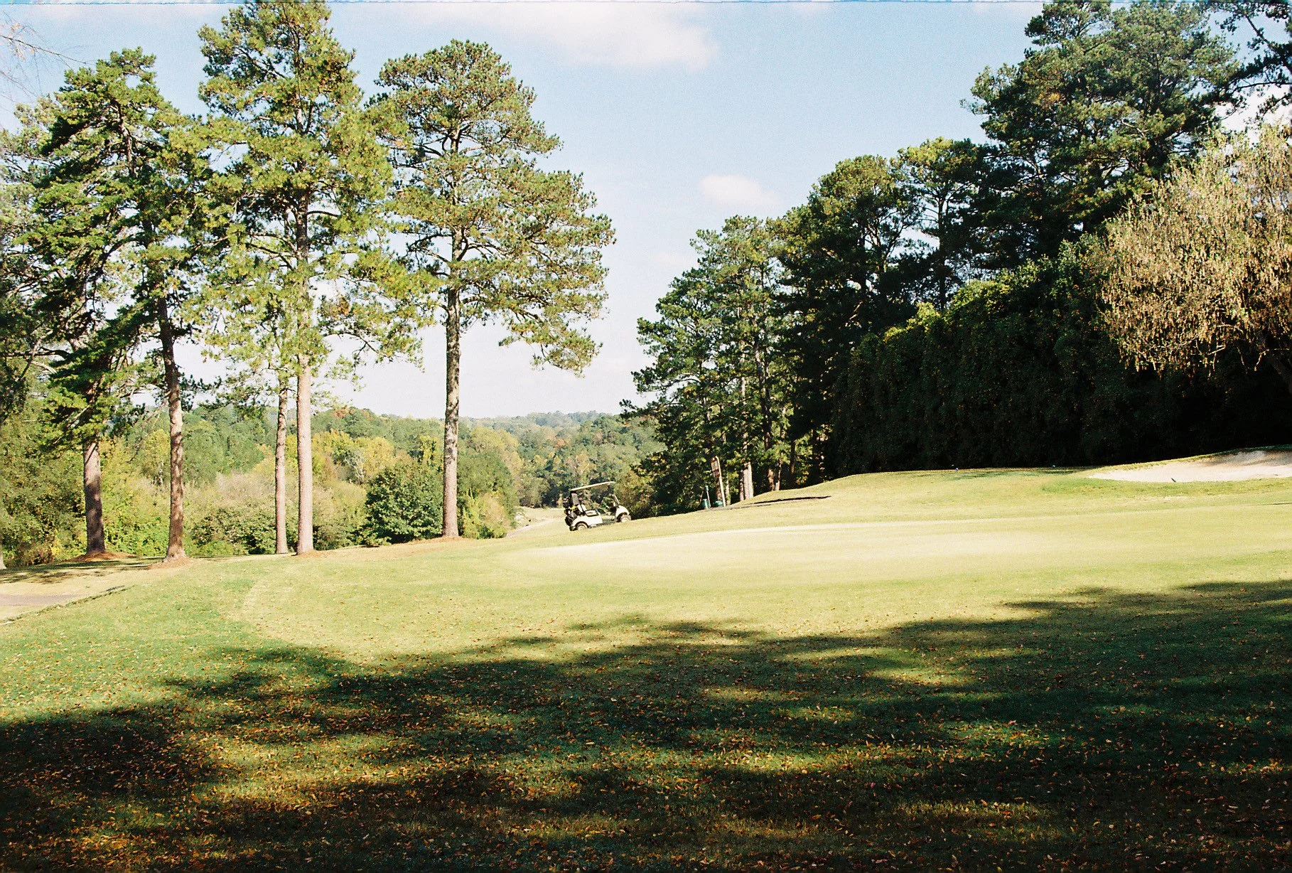 A golf course with a golf cart parked in the distance, surrounded by tall trees and green grass.