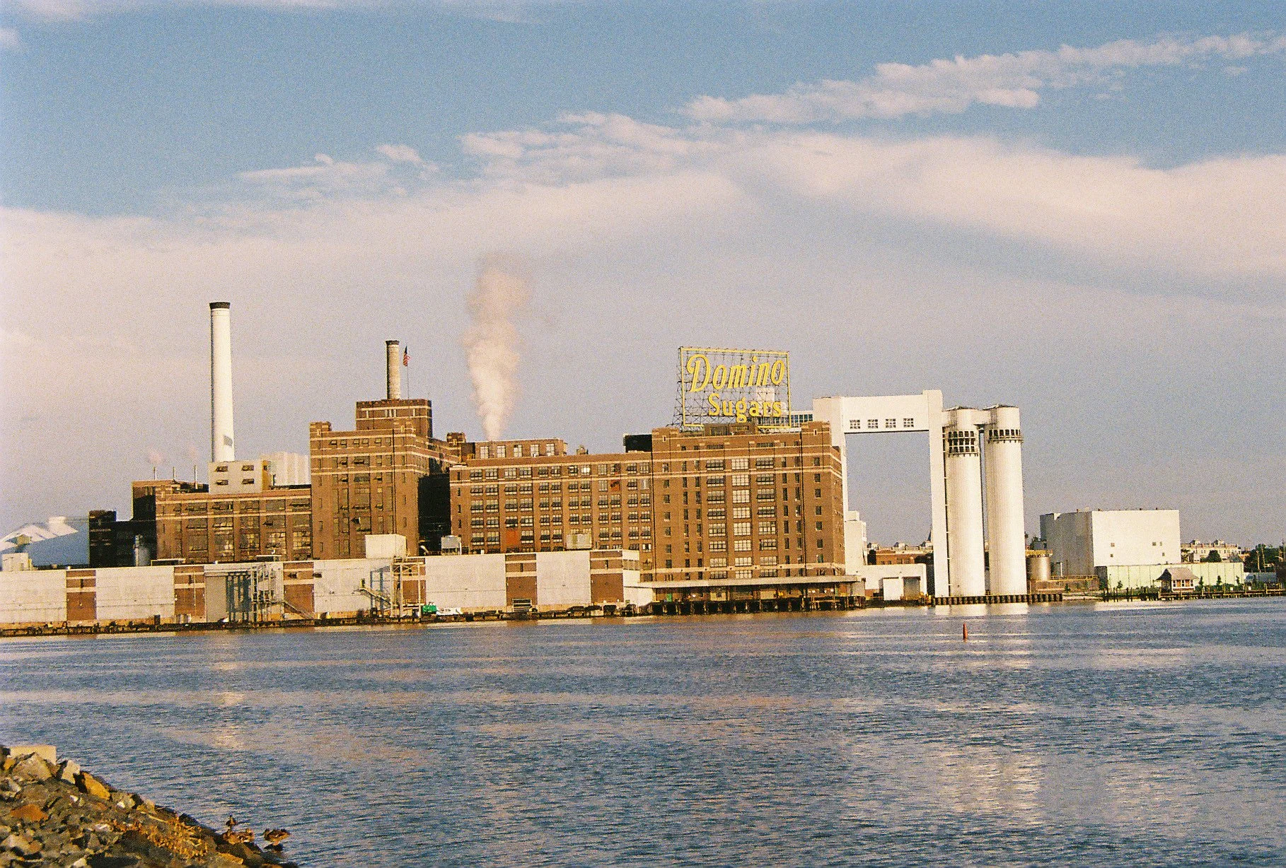 An industrial building by the water with a large yellow sign that reads 'Domino Sugars' on top, multiple smokestacks emitting smoke, and a cloudy sky in the background.