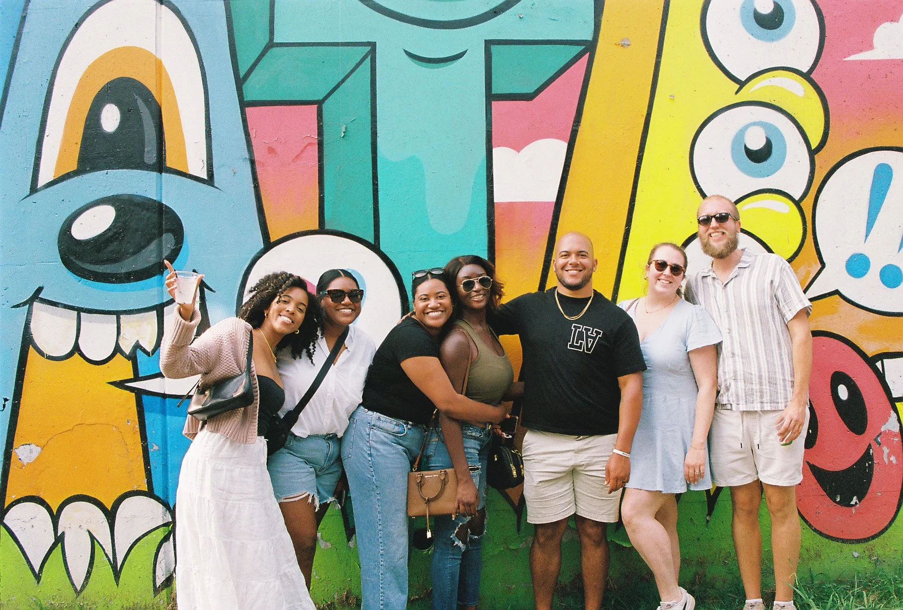 Group of seven diverse people smiling and posing in front of a colorful mural featuring cartoonish eyes, teeth, and abstract shapes.