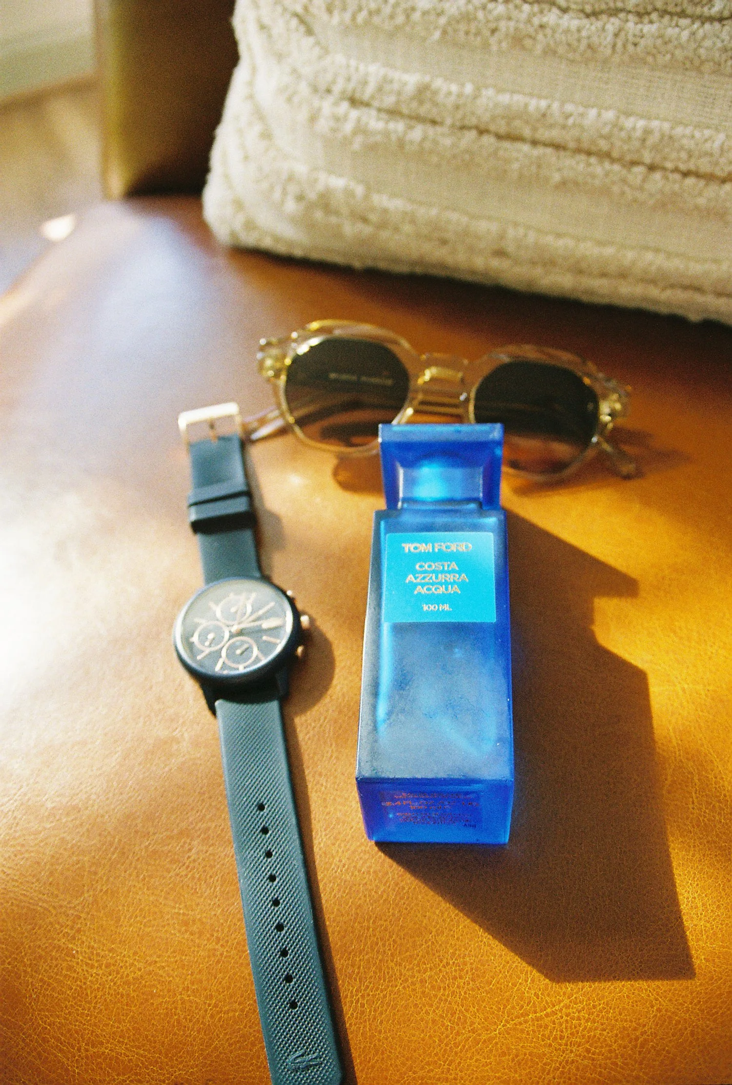 Sunglasses, a wristwatch, and a bottle of Tom Ford Costa Azzurra Acqua cologne on a leather surface with a towel in the background.