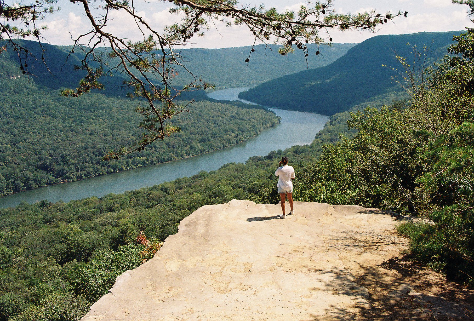 A person standing on a rocky ledge overlooking a lush green landscape with a winding river, framed by trees and mountains in the background.