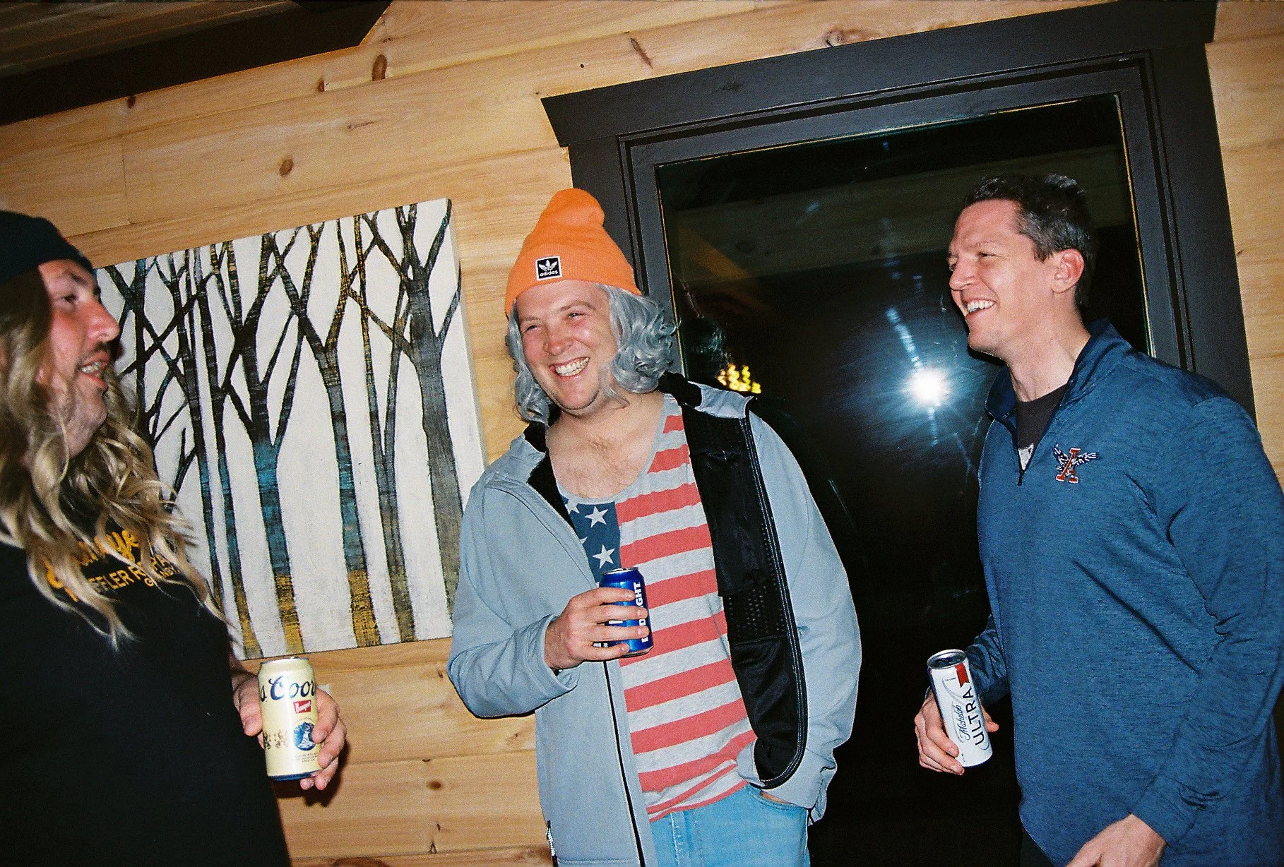 Three men with long hair, casual clothes, holding cans of beer, smiling and talking indoors in front of a wooden wall and a black framed window