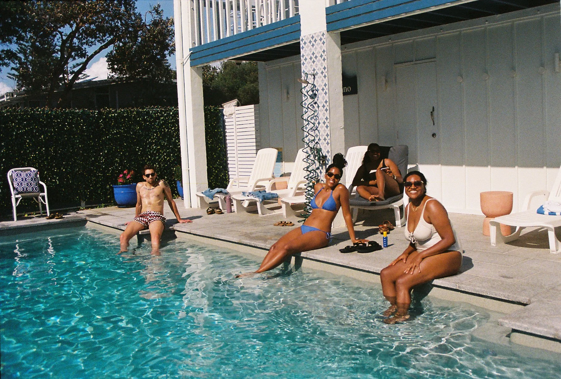 People relaxing by a swimming pool, sitting on the edge with their feet in the water. Four women are in the foreground, one man is in the background. They are wearing swimsuits and sunglasses. The poolside area has lounge chairs, potted plants, and a