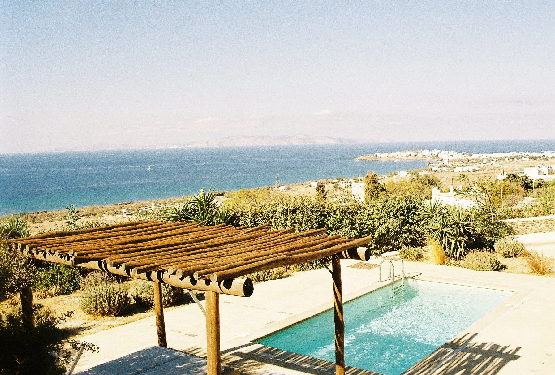 A swim pool with a wooden shade structure on a patio overlooking an ocean view with distant land, green trees, shrubs, and vegetation in the foreground.