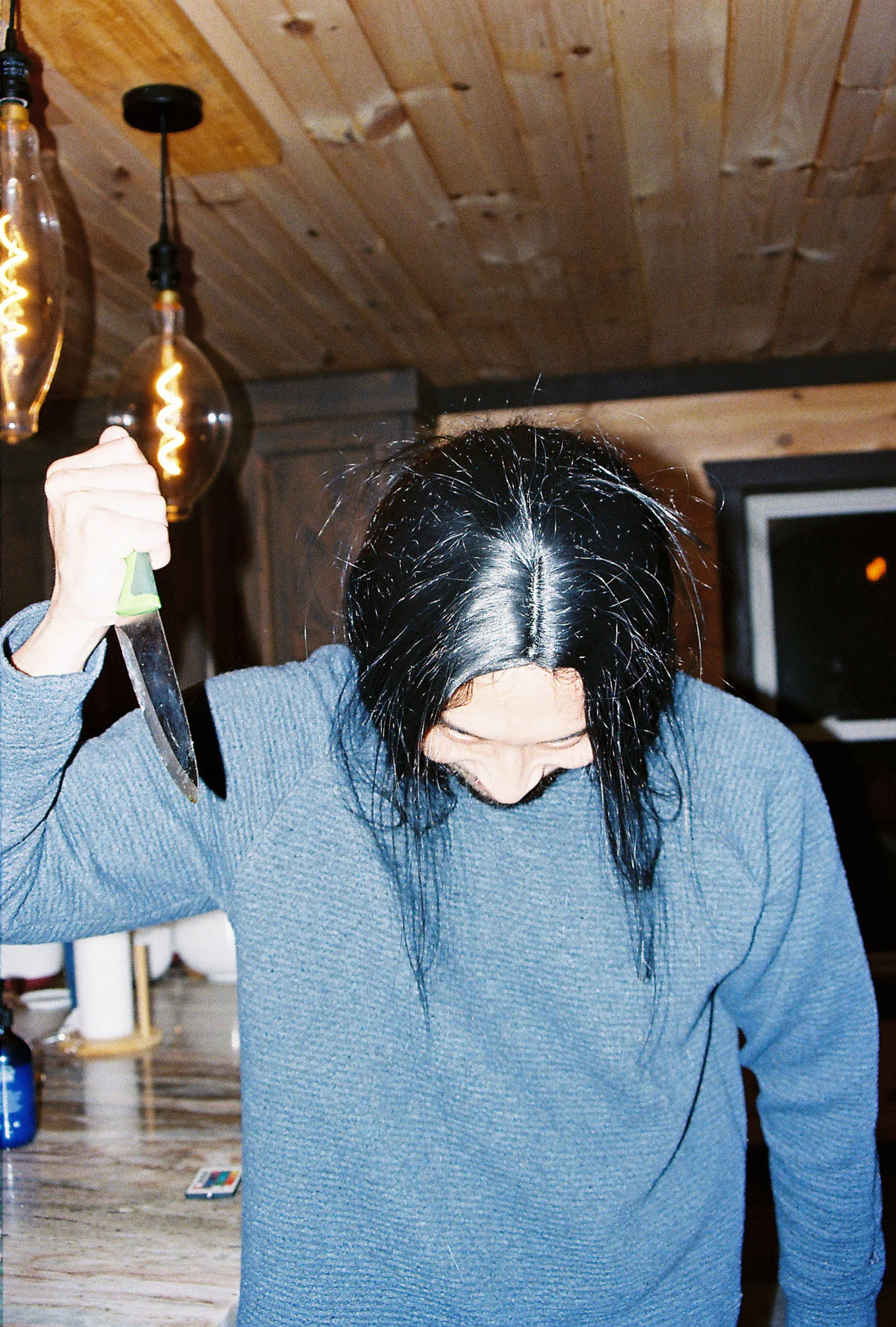 Person with black hair and a gray sweater holding a kitchen knife, looking down, in a room with wooden ceiling and hanging light bulbs.