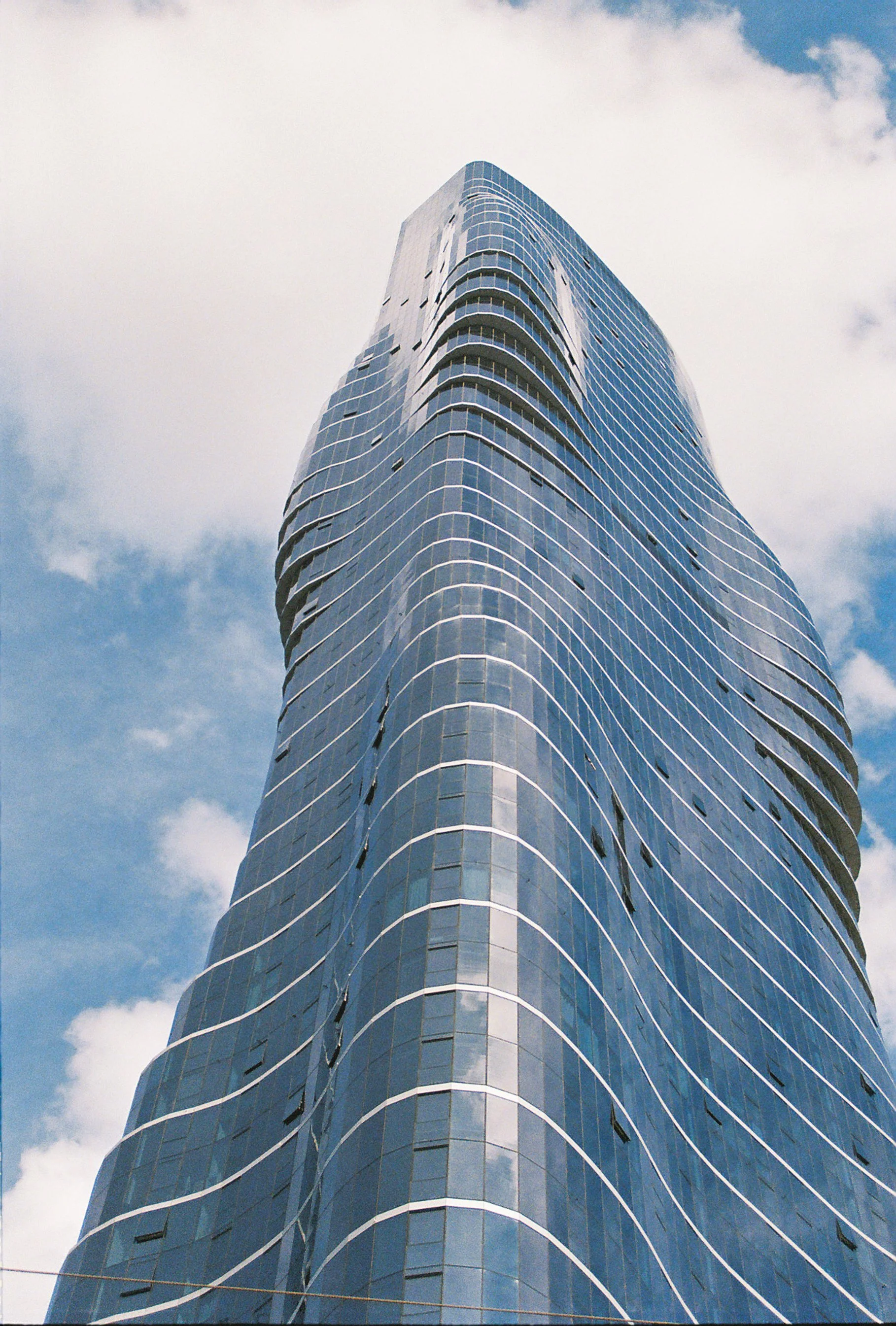 A tall modern glass skyscraper viewed from the ground up, reflecting the partly cloudy sky.