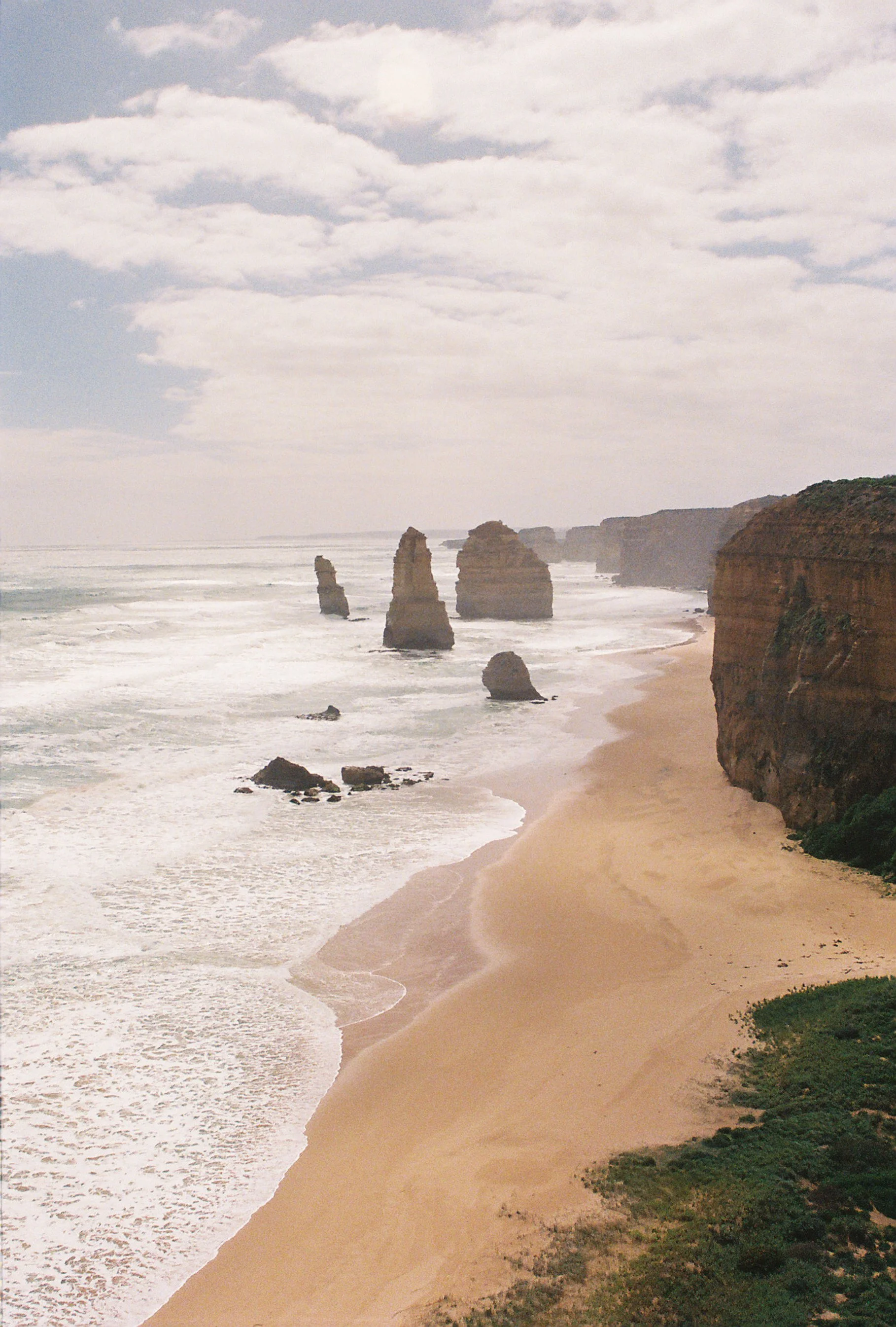 View of a sandy beach with four large rock formations in the ocean, cliffs on the right side, and partly cloudy sky overhead.