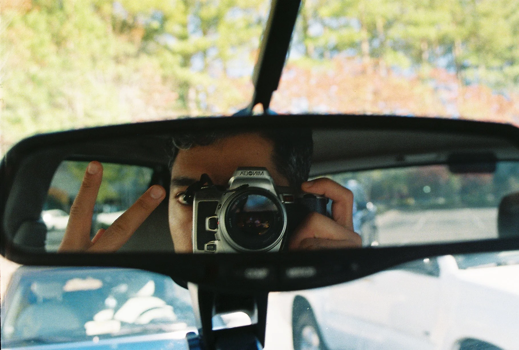 Person taking a photo with a camera in a car's rearview mirror, holding up peace sign, with trees and parking lot outside.