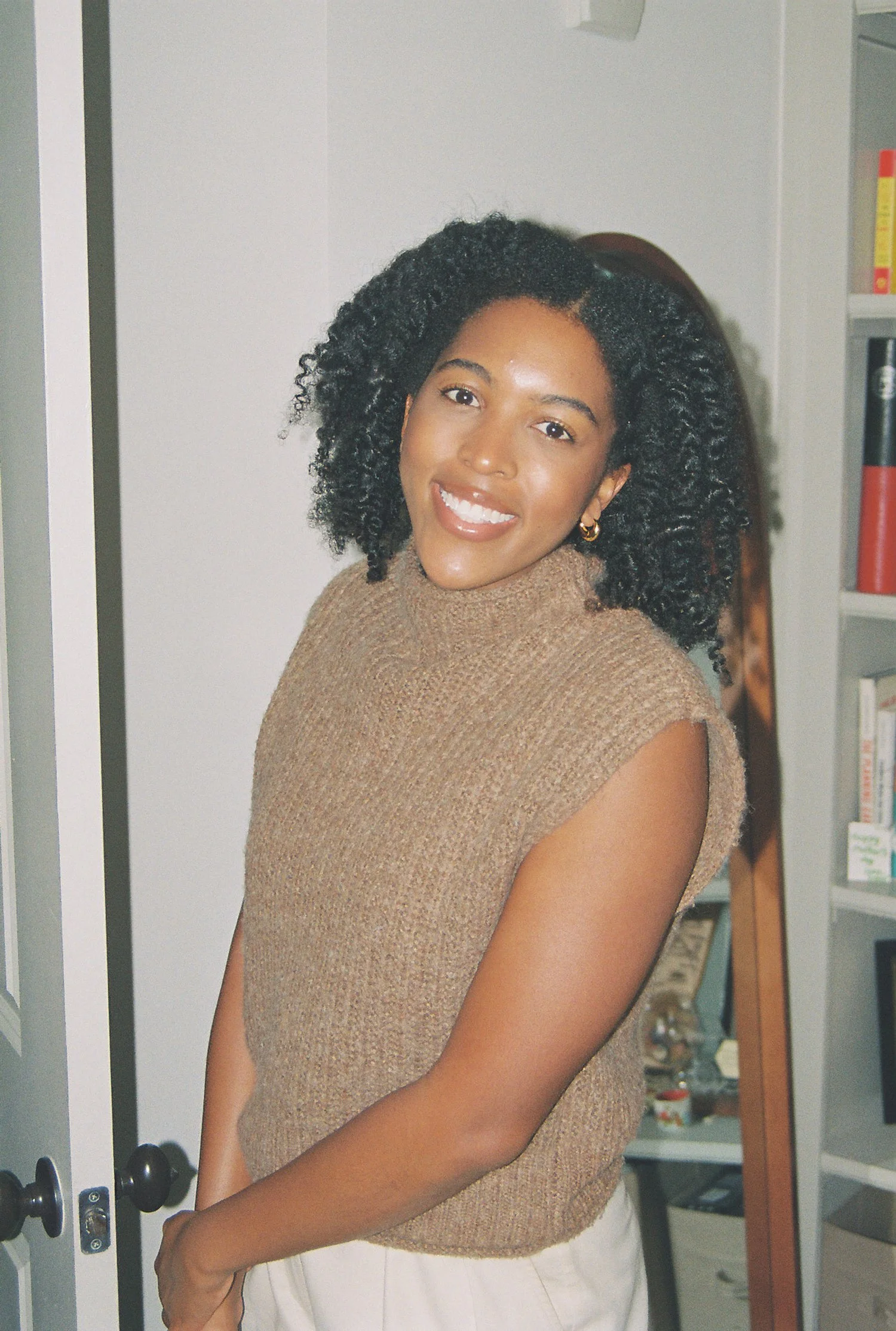 A smiling woman with curly hair wearing a brown sleeveless turtleneck top, standing in front of a bookshelf filled with books.