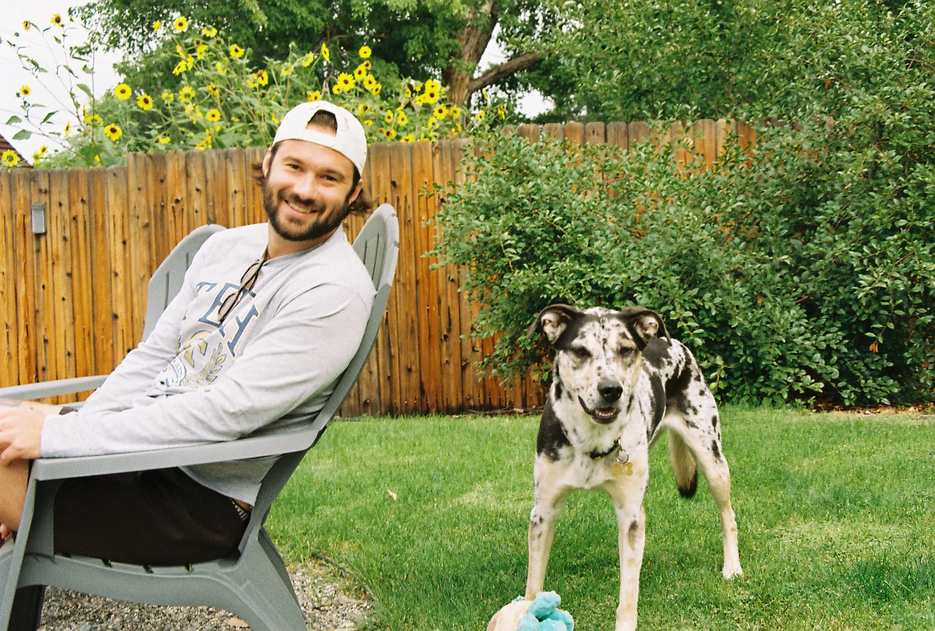 A smiling young man with a beard sitting on a gray outdoor chair in a backyard, wearing a white backwards cap and a gray long-sleeve shirt. Next to him, a black and white speckled dog stands on the grass, looking at the camera. In the background, the