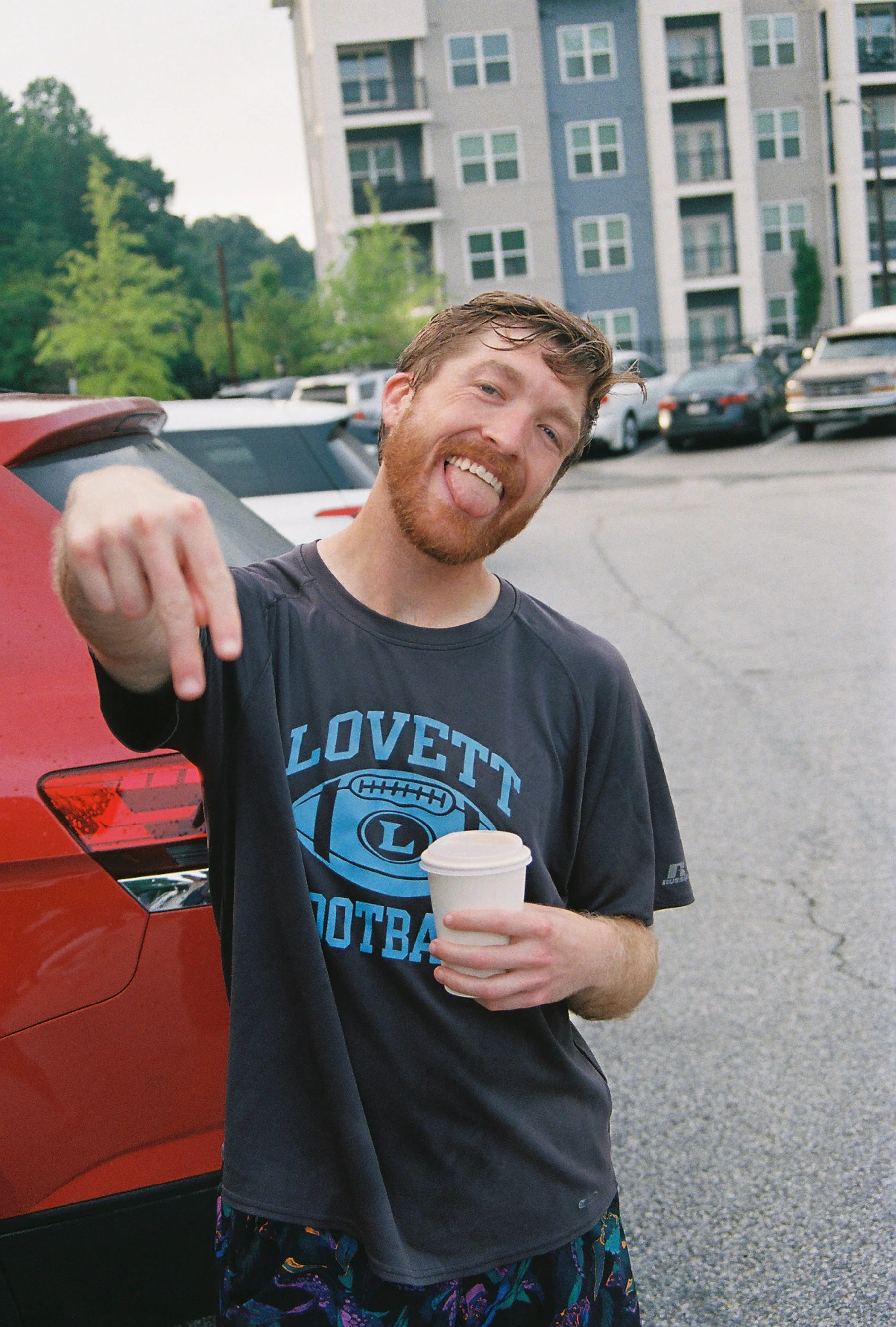 A smiling man with a beard and wet hair sticking out his tongue, wearing a Lovett Football t-shirt, holding a coffee cup, standing outdoors near a red car in a parking lot with condo buildings in the background.
