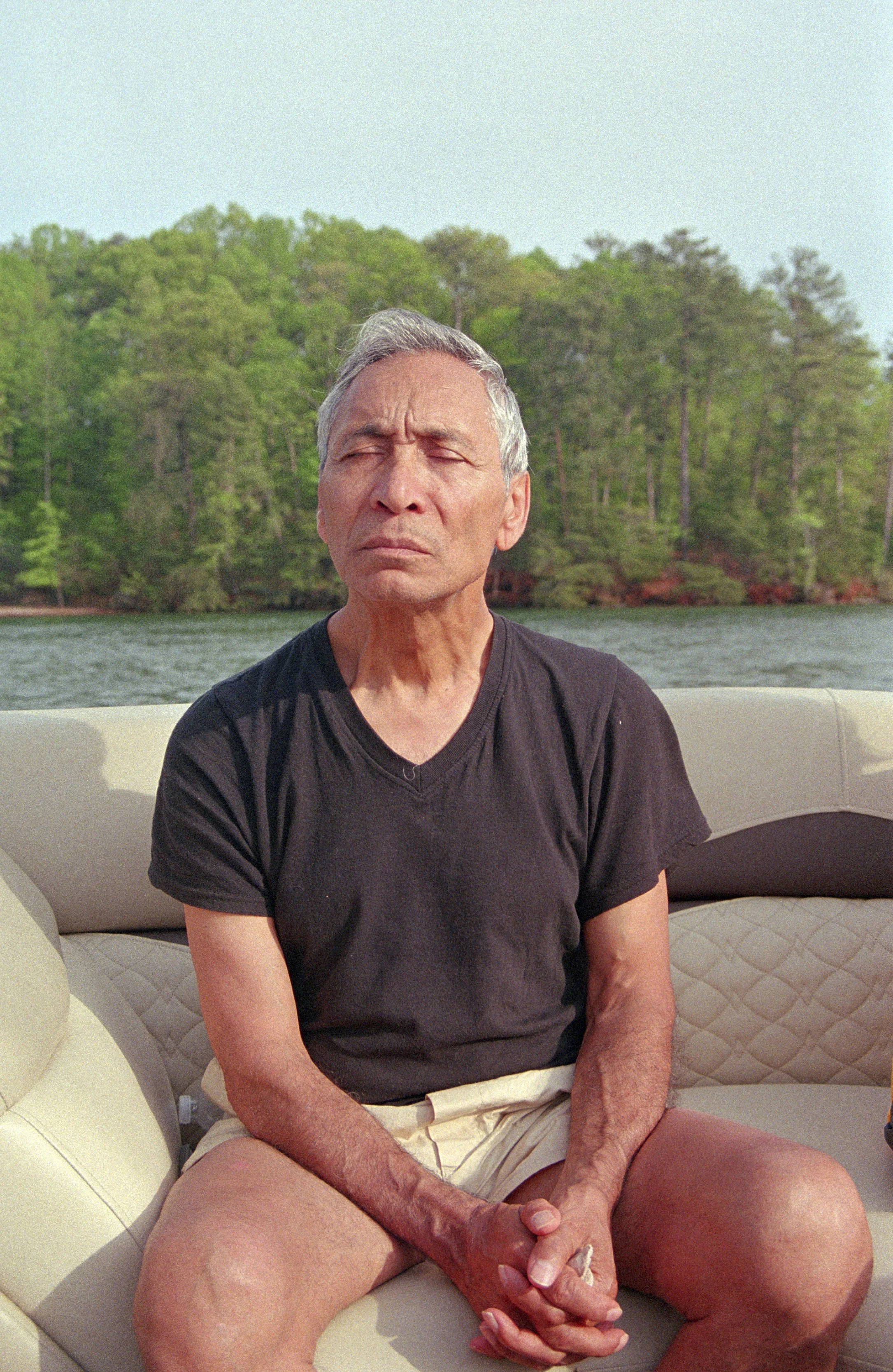 An elderly man with gray hair sitting on a boat with trees and water in the background, wearing a black t-shirt and beige shorts, with closed eyes and a serious expression.