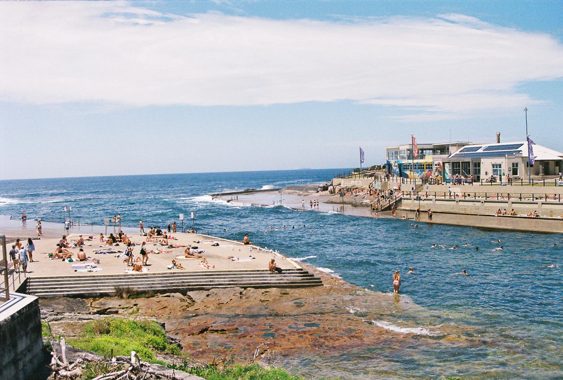 Beach scene with people sunbathing on a concrete area and swimming in the ocean. There are steps leading down to the rocky shoreline, and a building on a raised platform with flags on it.