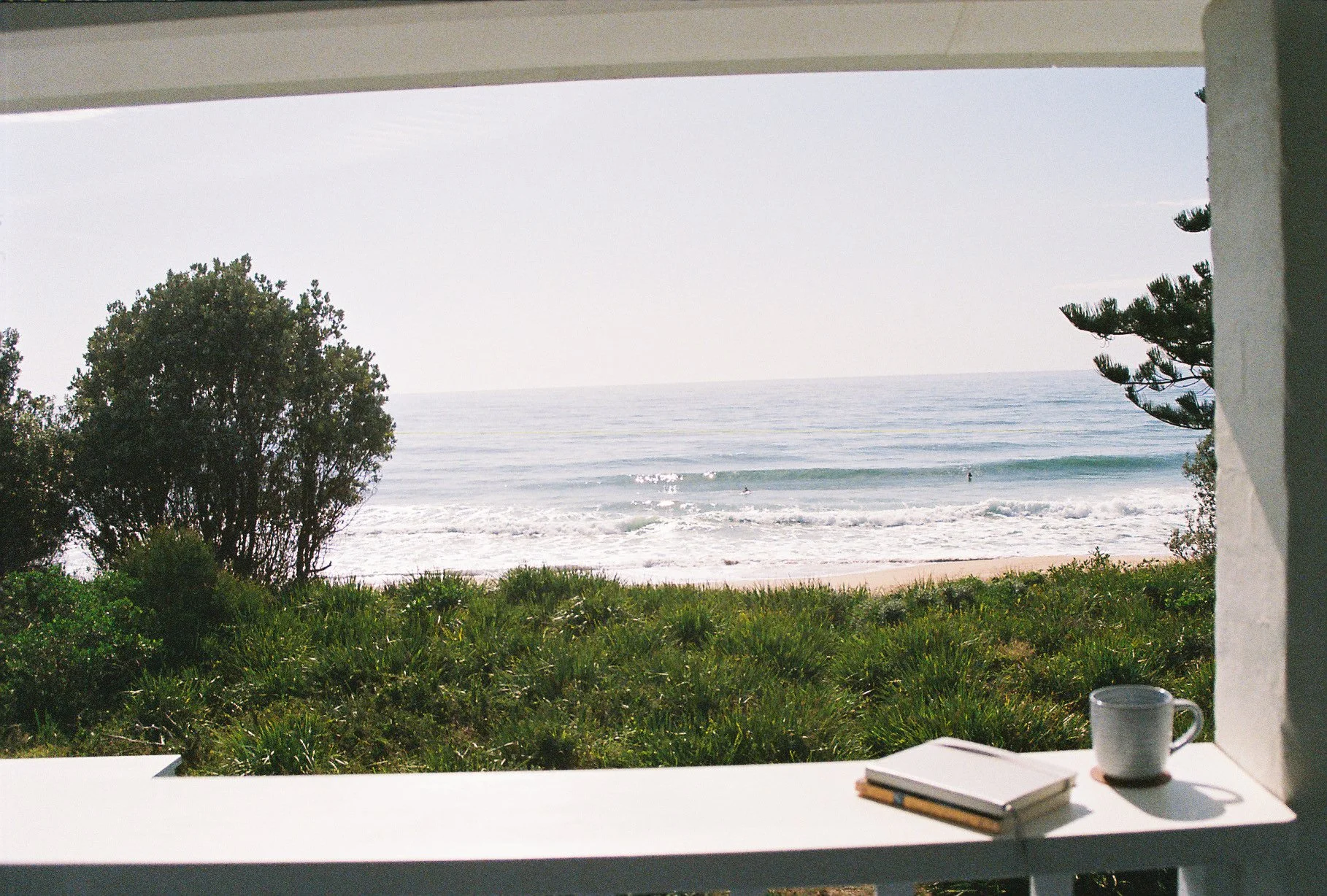 View of the beach and ocean through a window, with a white shelf in the foreground holding books and a cup.