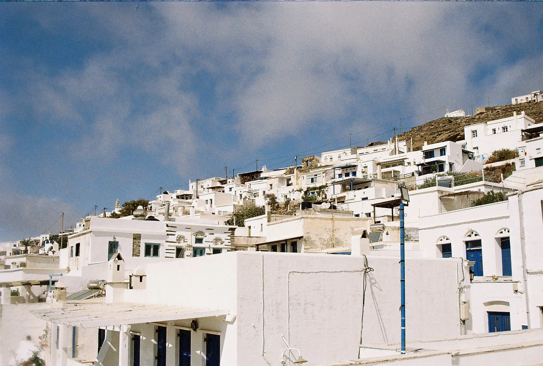 White buildings with blue accents on a hillside against a partly cloudy sky, typical of Cycladic architecture.