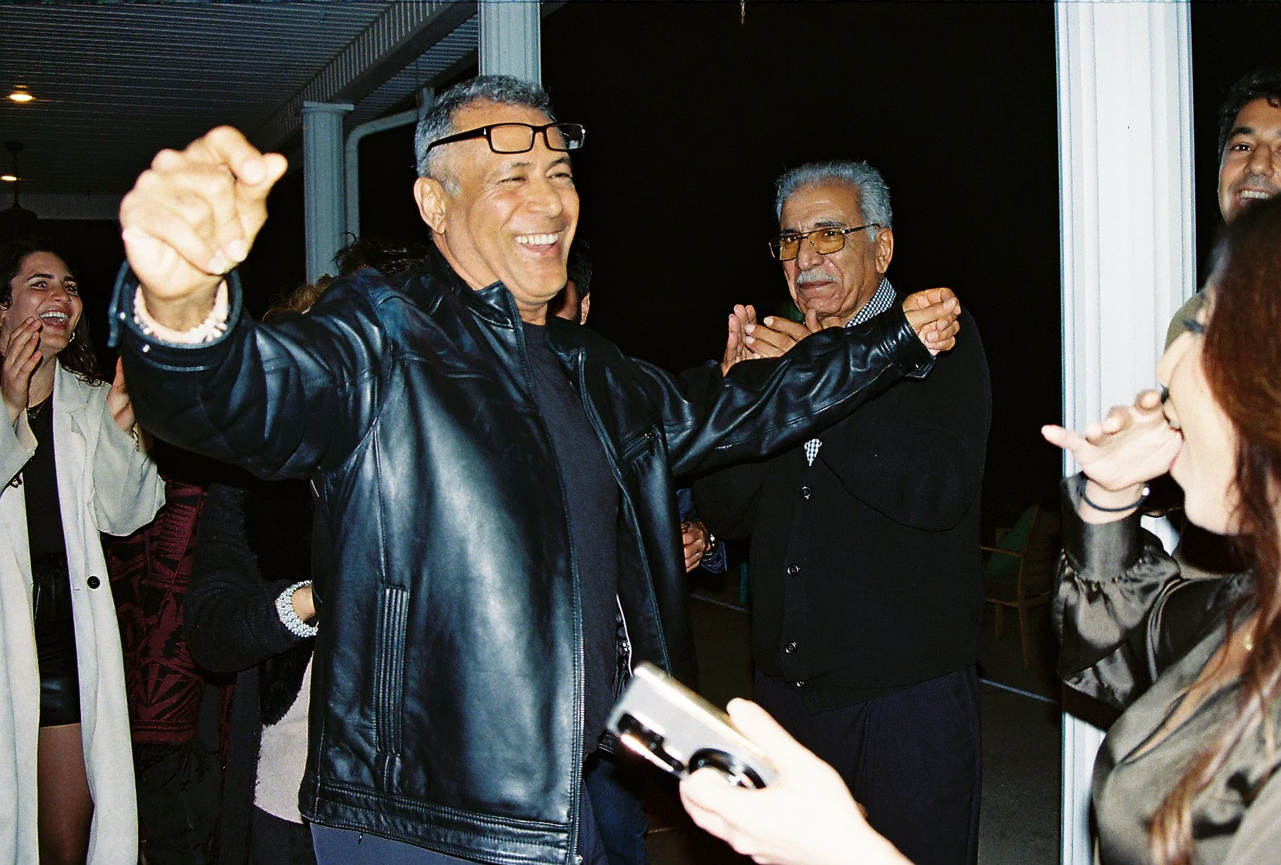 A group of people celebrating, with a man in a black leather jacket smiling and extending his arms, surrounded by others in a social setting.