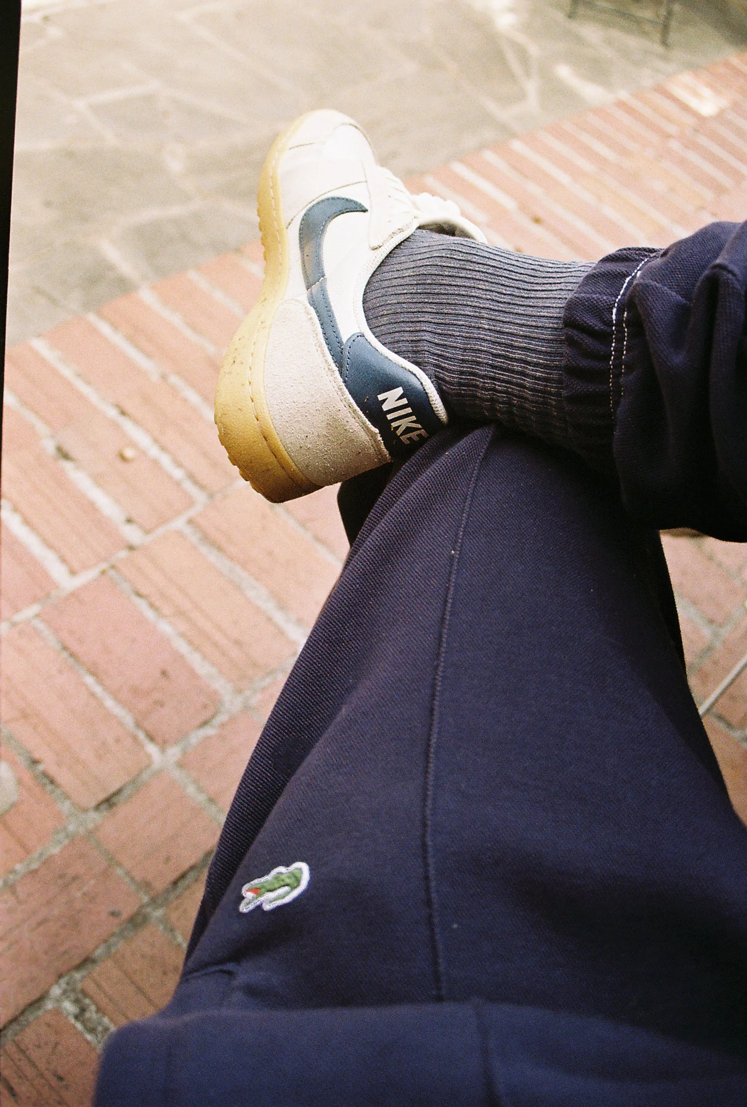 Person sitting outdoors with legs crossed, wearing dark pants with a Lacoste logo, Nike sneakers, and dark socks, on a brick and concrete surface.
