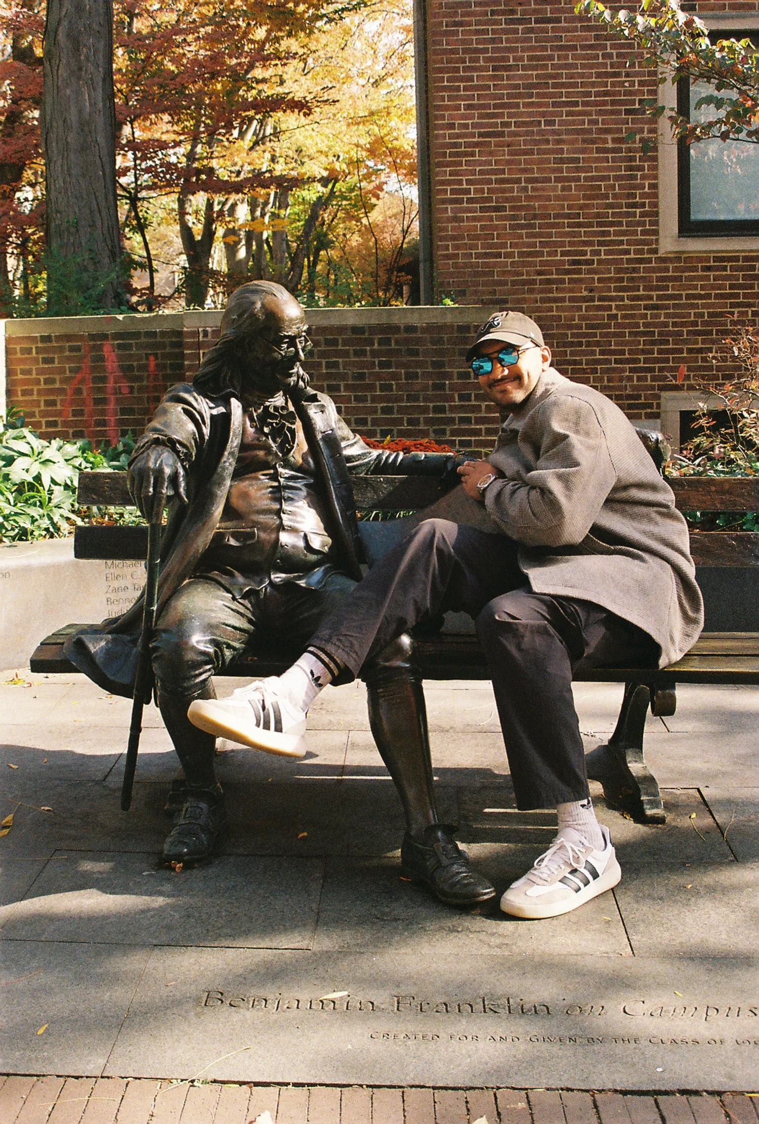 A man sitting on a park bench next to a bronze statue of Benjamin Franklin, which shows Franklin leaning back and holding a cane. The man is smiling, wearing sunglasses, a gray coat, black pants, white sneakers with black stripes, and a cap. The scen