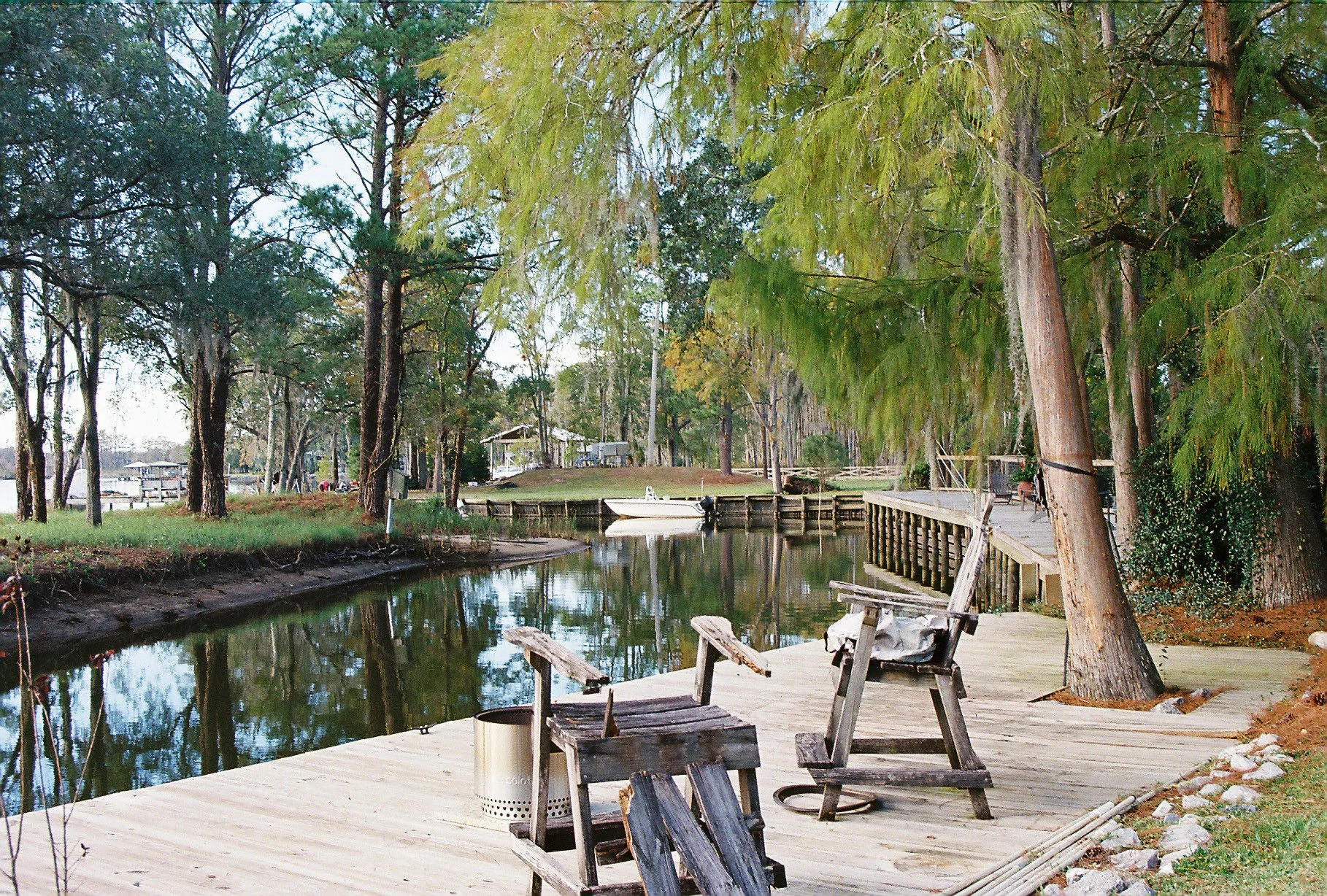 A peaceful riverside scene with a wooden dock, empty wooden chairs, a small boat, and tall trees with green foliage.