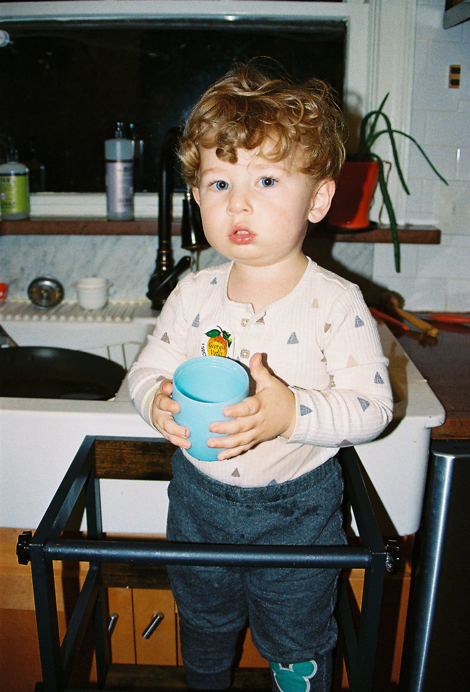 A young boy with curly hair holding a blue cup, standing on a black kitchen table in a kitchen with a sink, counter, and potted plant in the background.