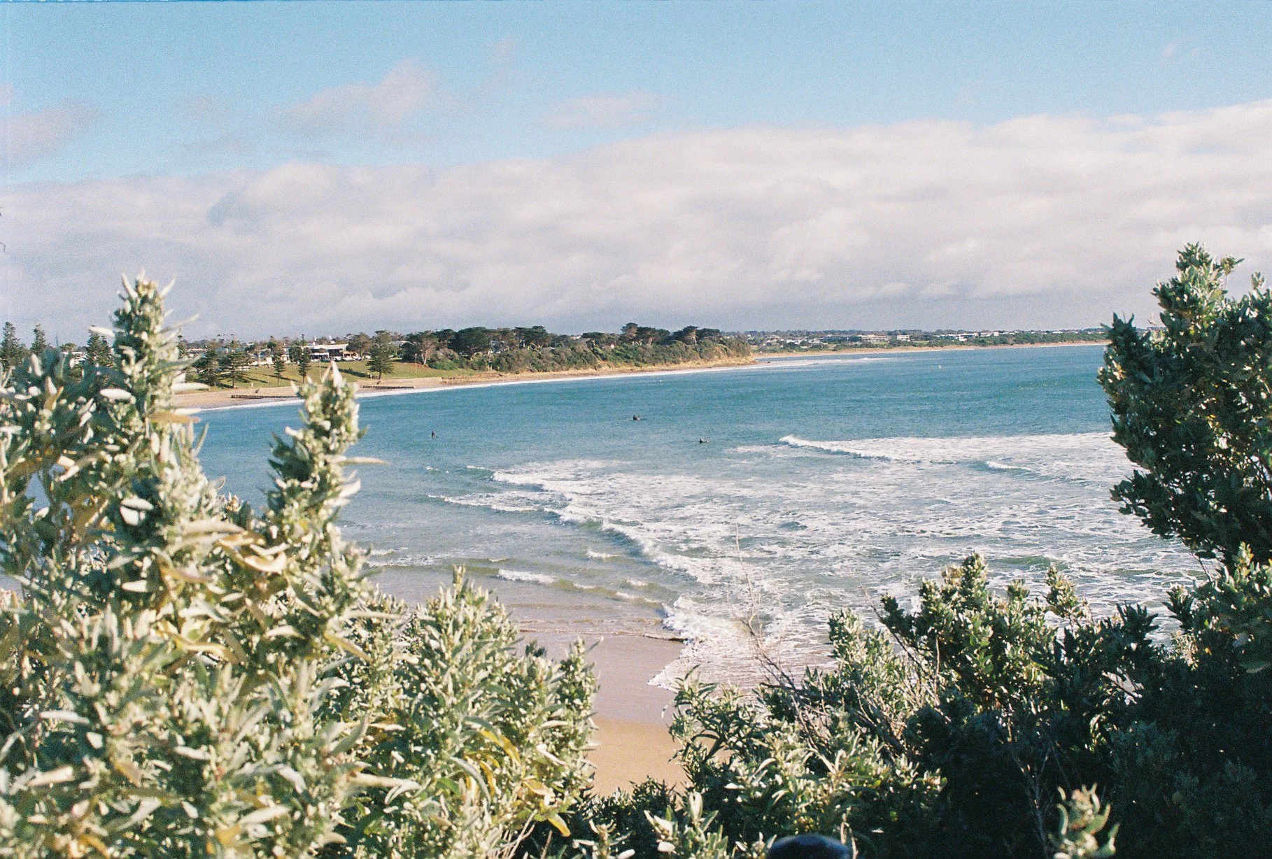 View of a beach with ocean waves, sandy shore, and greenery in the foreground, under a partly cloudy sky.
