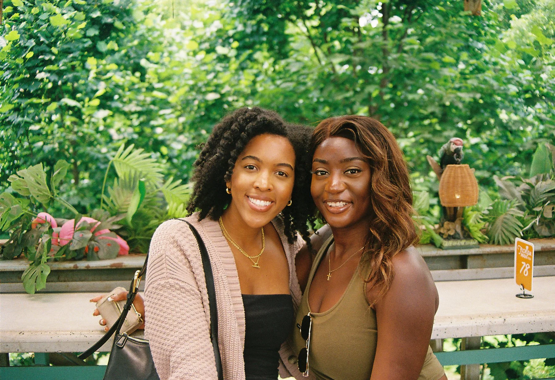 Two women smiling and standing close together in a lush green indoor garden or botanical setting.