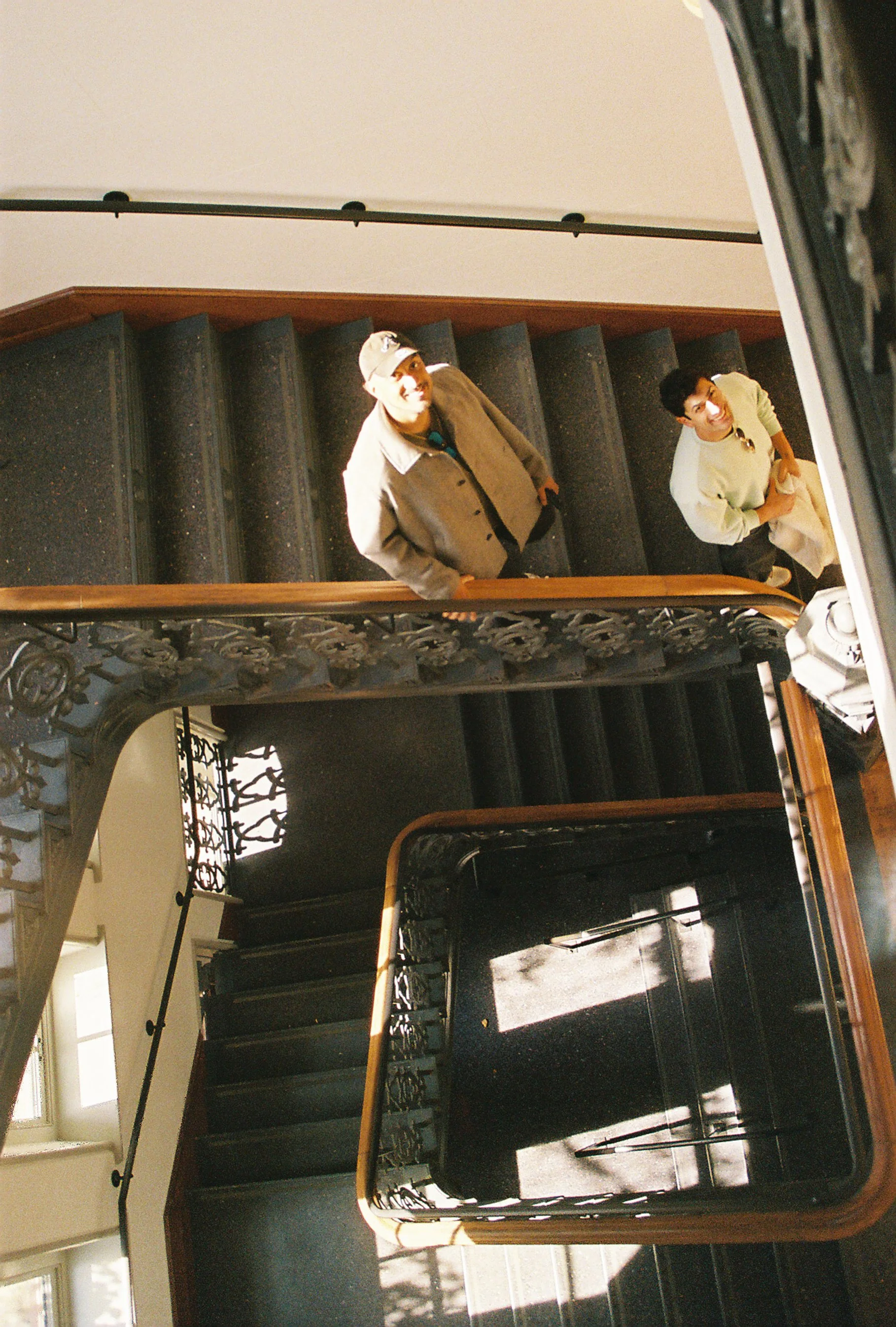 Two people standing on a black carpeted staircase, looking up at the camera, with ornate black iron railings and a wooden handrail.