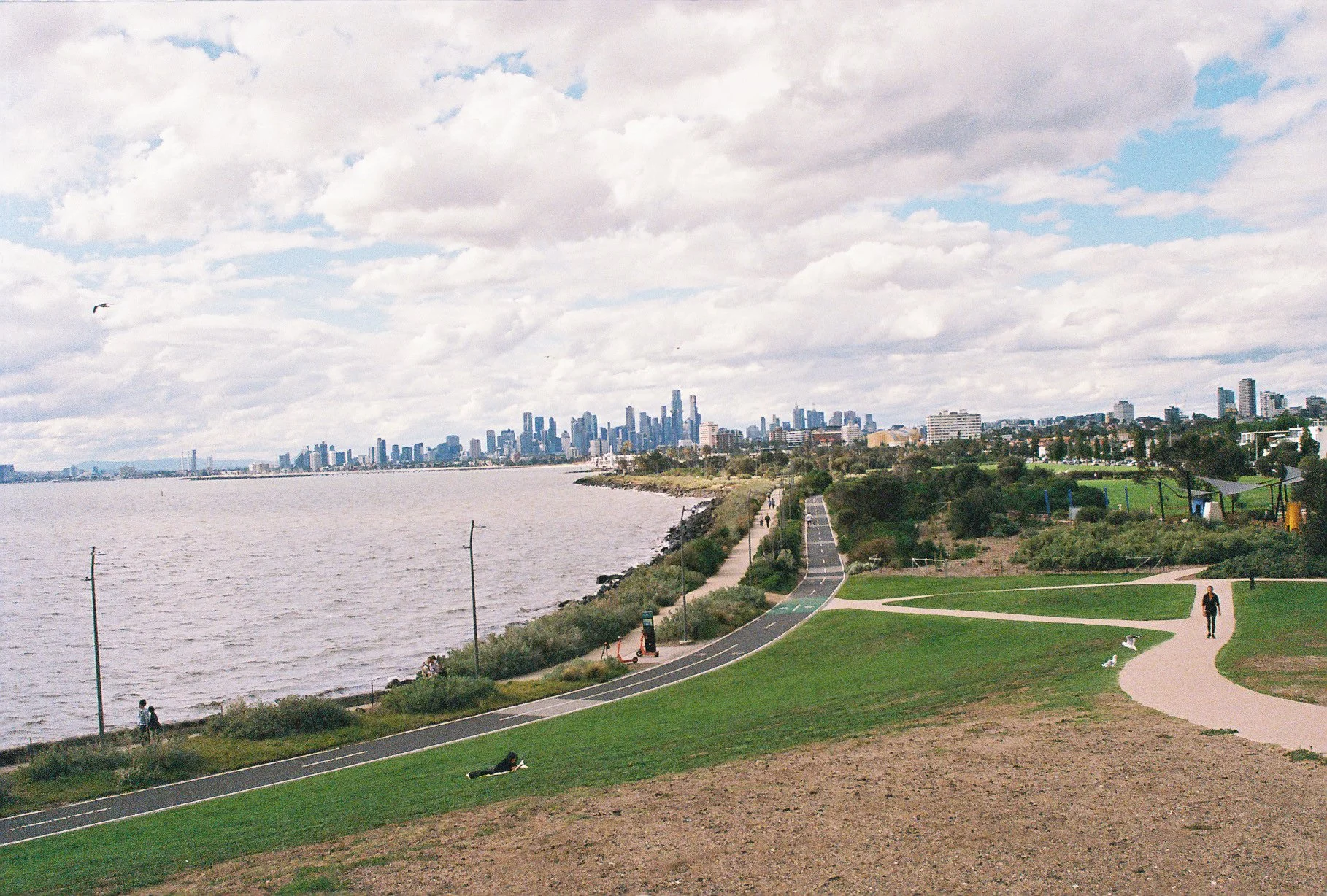View of a city skyline across a body of water with a park and walking paths in the foreground, some people walking and relaxing, under a partly cloudy sky.