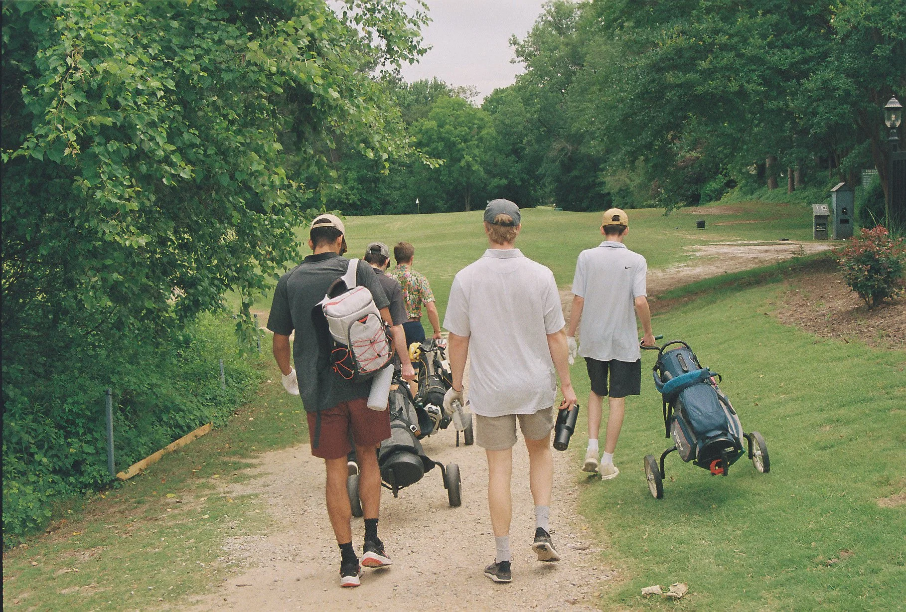 Four people walking on a golf course path with golf bags and push carts, surrounded by trees and greenery.