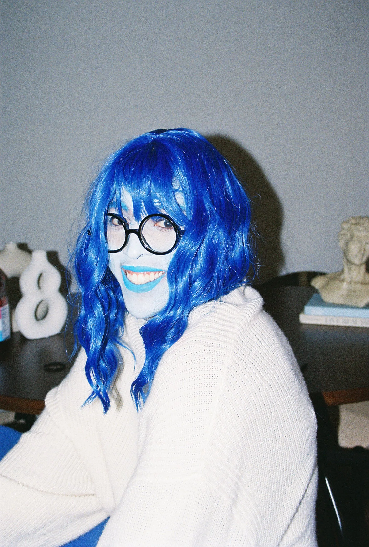 Person with bright blue wavy hair, wearing black round glasses, smiling with blue lipstick, dressed in a cream-colored sweater, sitting at a desk with art supplies and decor in the background.