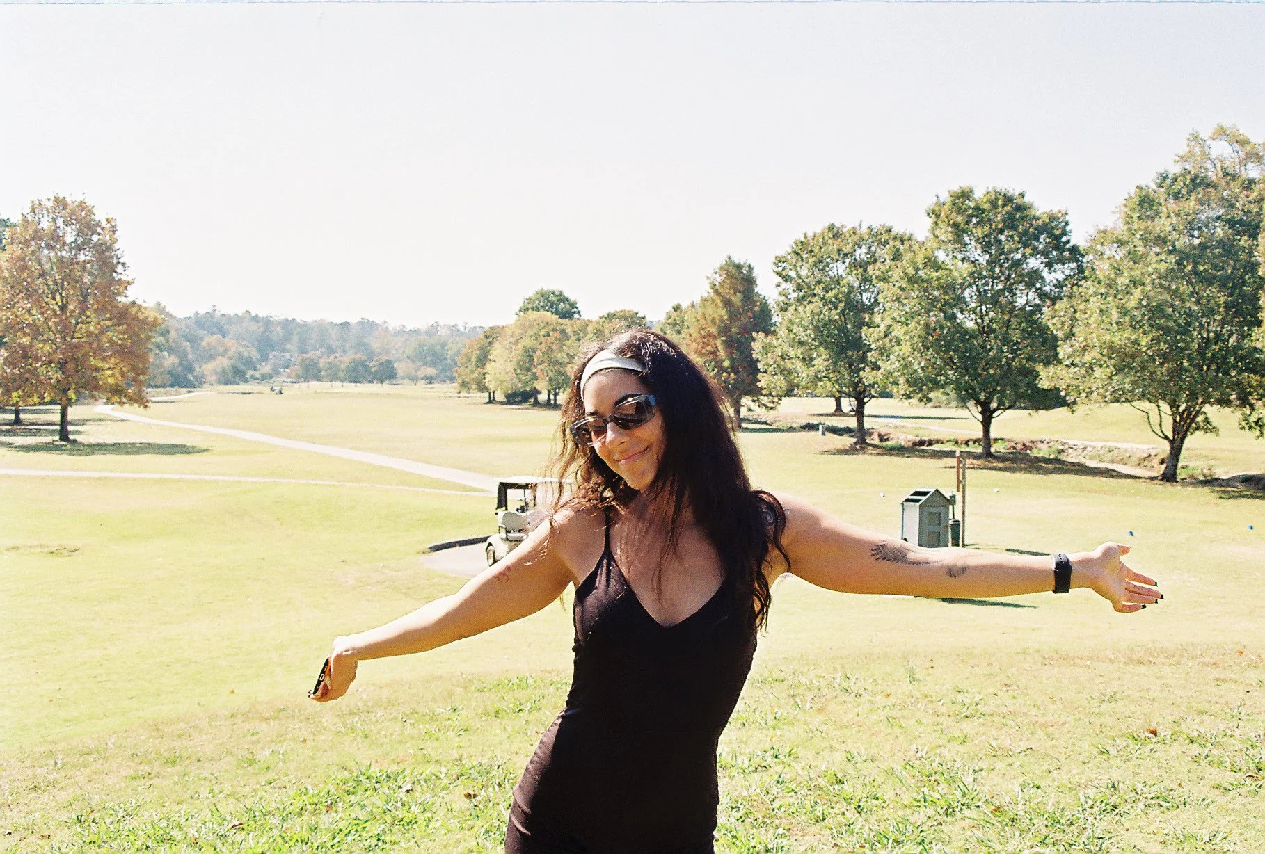 A woman in sunglasses and a black dress standing outdoors on a sunny day with outstretched arms, smiling, in a park with trees and golf carts in the background.