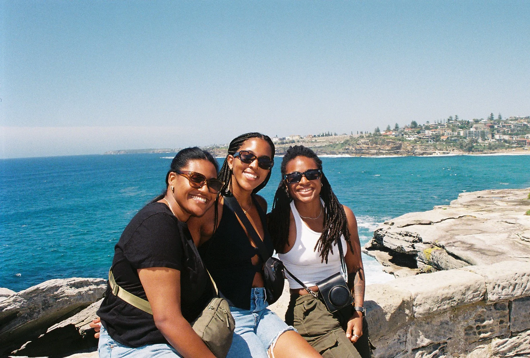 Three women smiling, wearing sunglasses, sitting on rocks with the ocean and a coastline in the background.