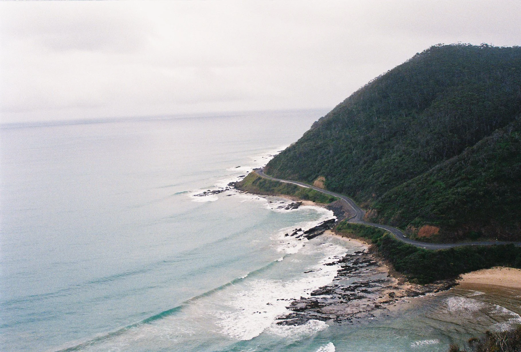 Aerial view of a coastal landscape with a winding road along the edge of a lush green hill and the ocean waves crashing onto the shore under an overcast sky.